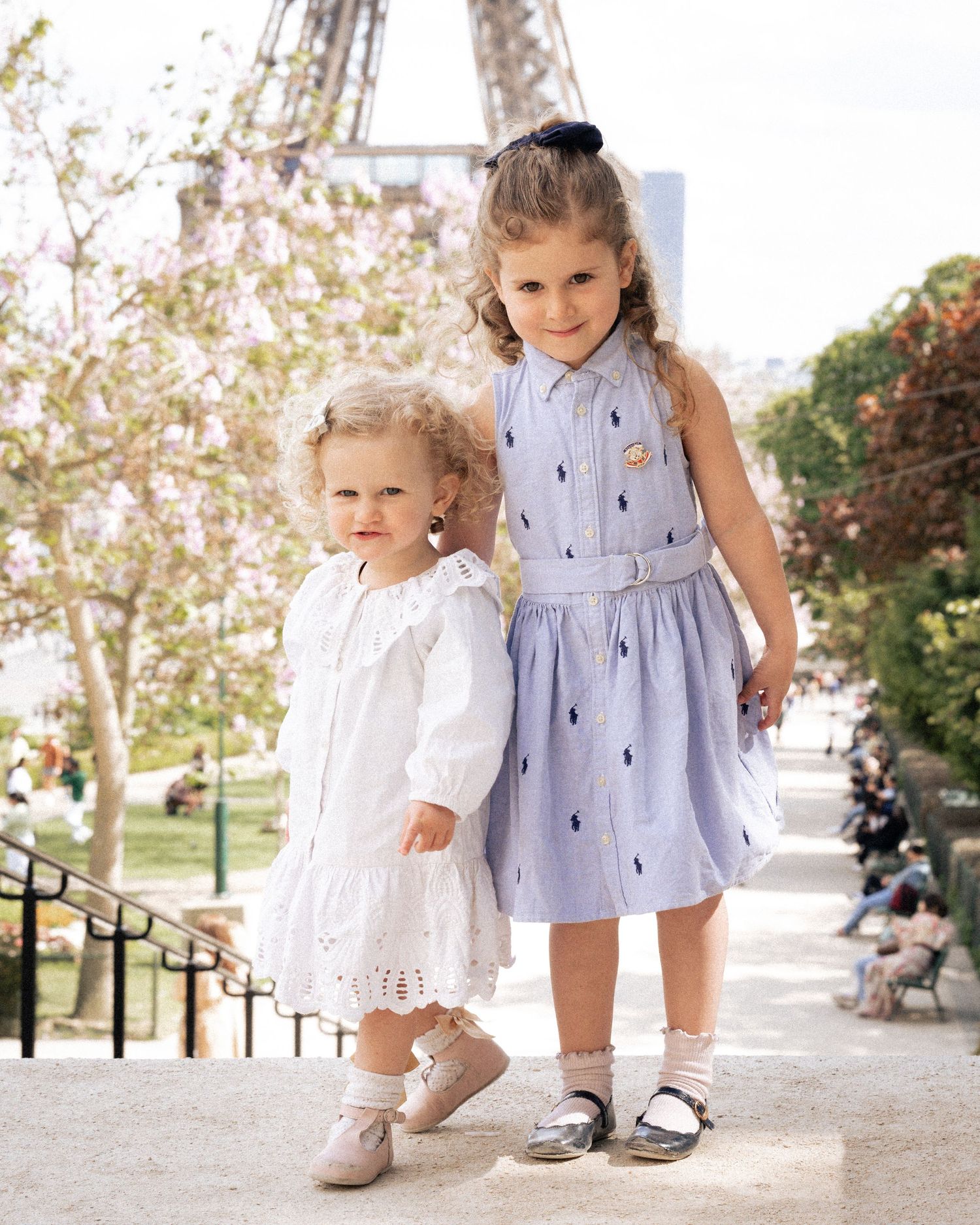 Two young girls in elegant dresses posing near Eiffel Tower in Paris on sunny day.