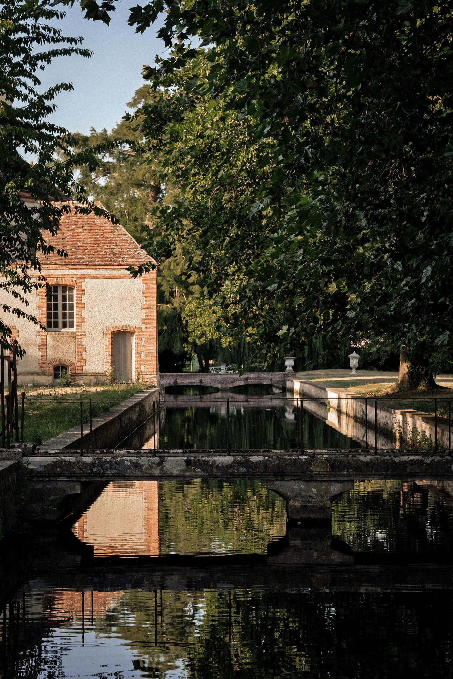 Historic brick building reflected in a calm moat surrounded by lush green trees in France.