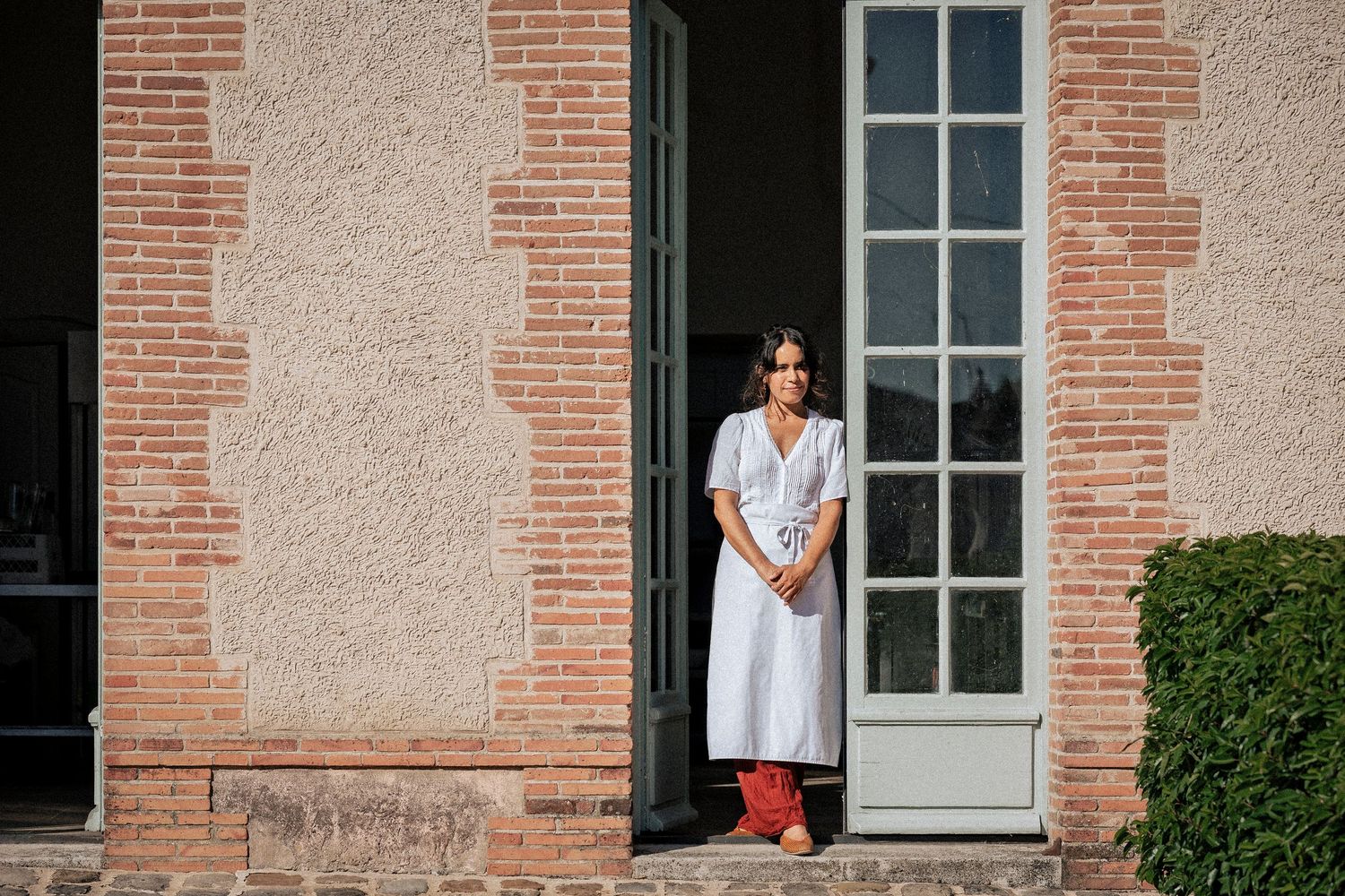 A woman in a white dress stands elegantly in a doorway of a rustic brick building.