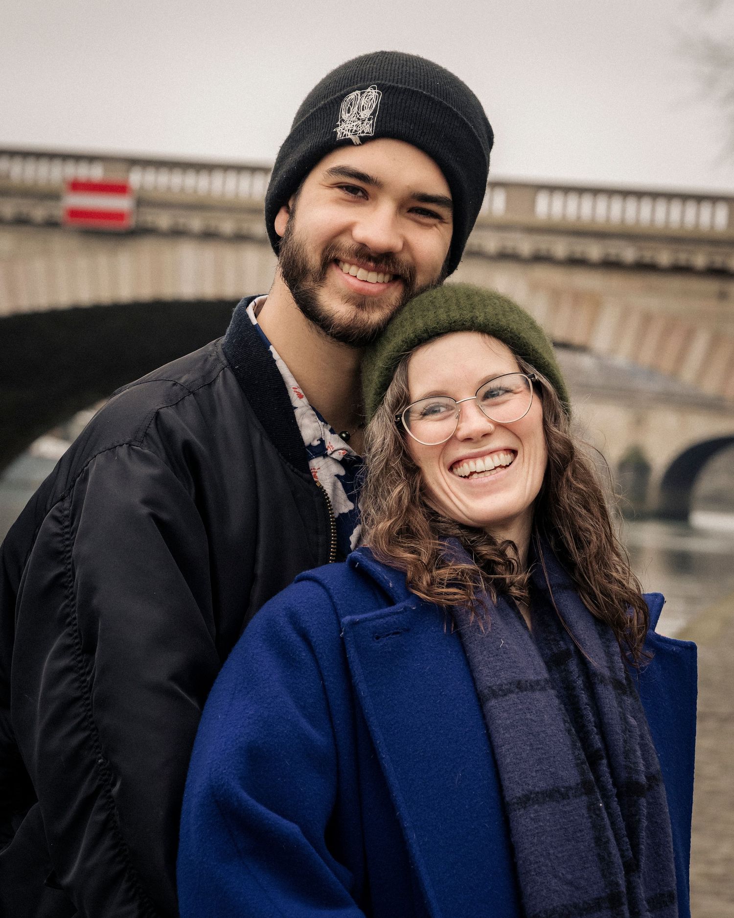 Happy young couple smiling together outdoors near a stone bridge on a cloudy winter day.