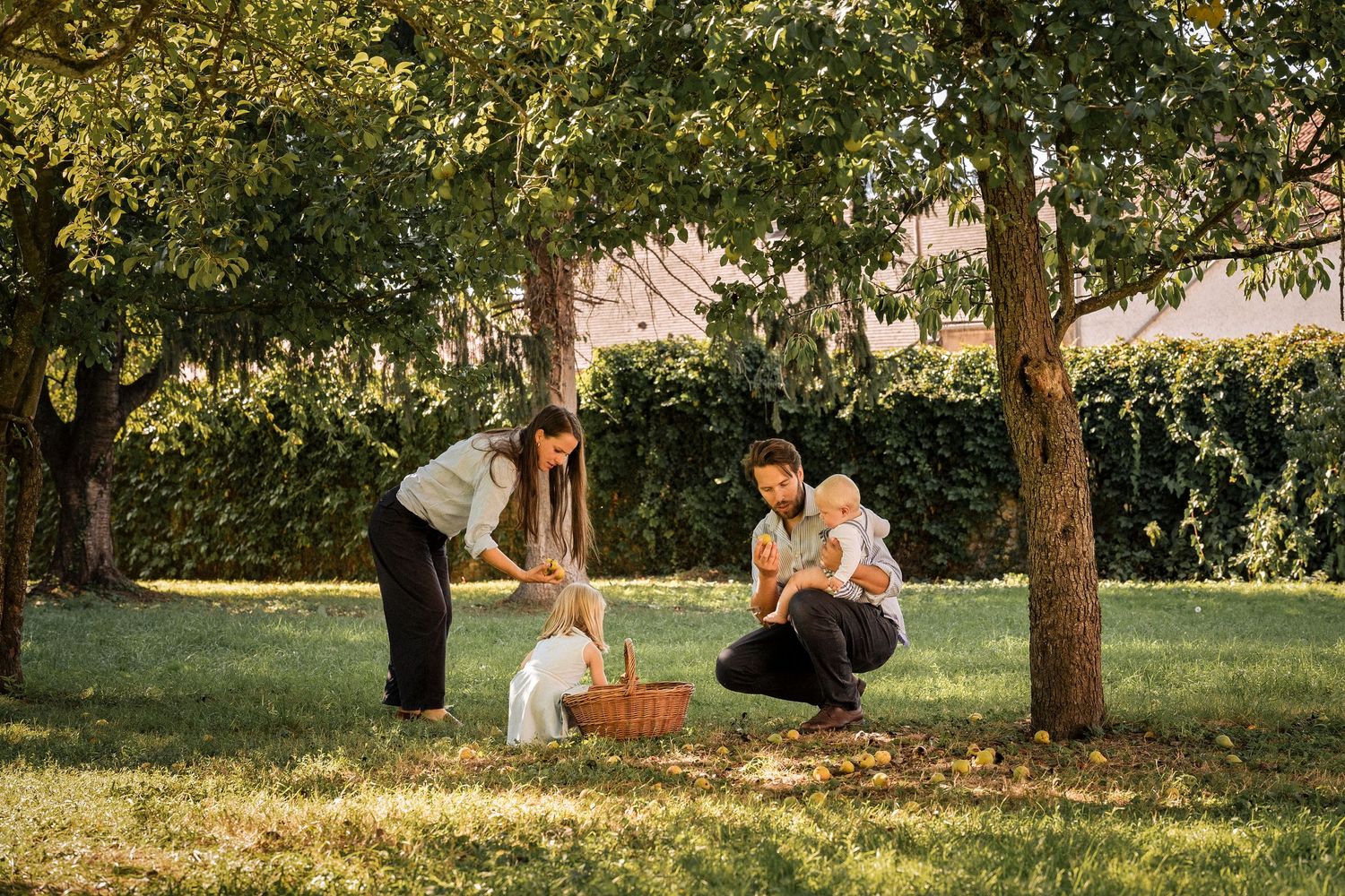 Family with two young children enjoying a sunny picnic in a lush green garden with trees.