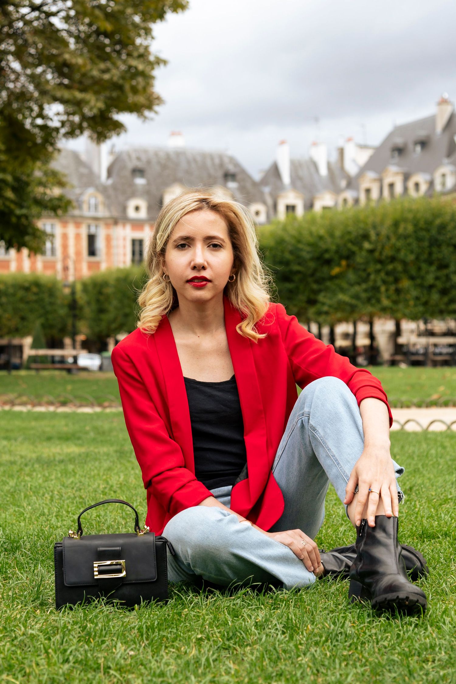 Blonde woman in red blazer sitting on grass in park with black handbag.