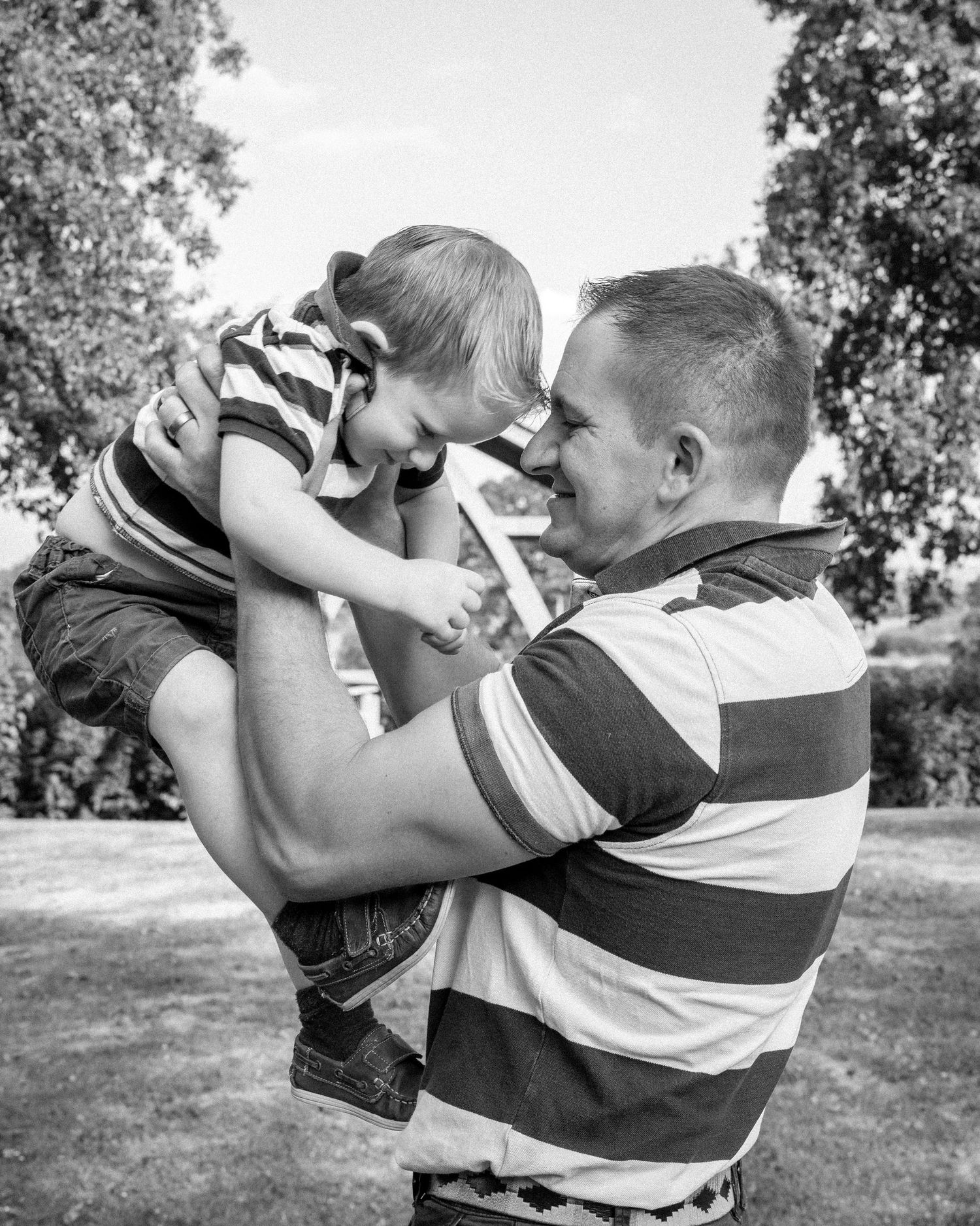 Black and white photo of a smiling man lifting a laughing toddler outdoors with trees in background.