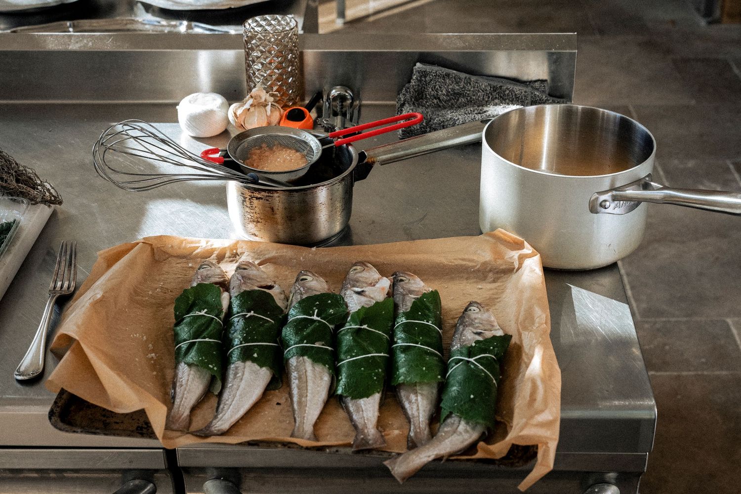 Fresh fish fillets wrapped in green leaves on parchment paper in a professional kitchen with cooking utensils.