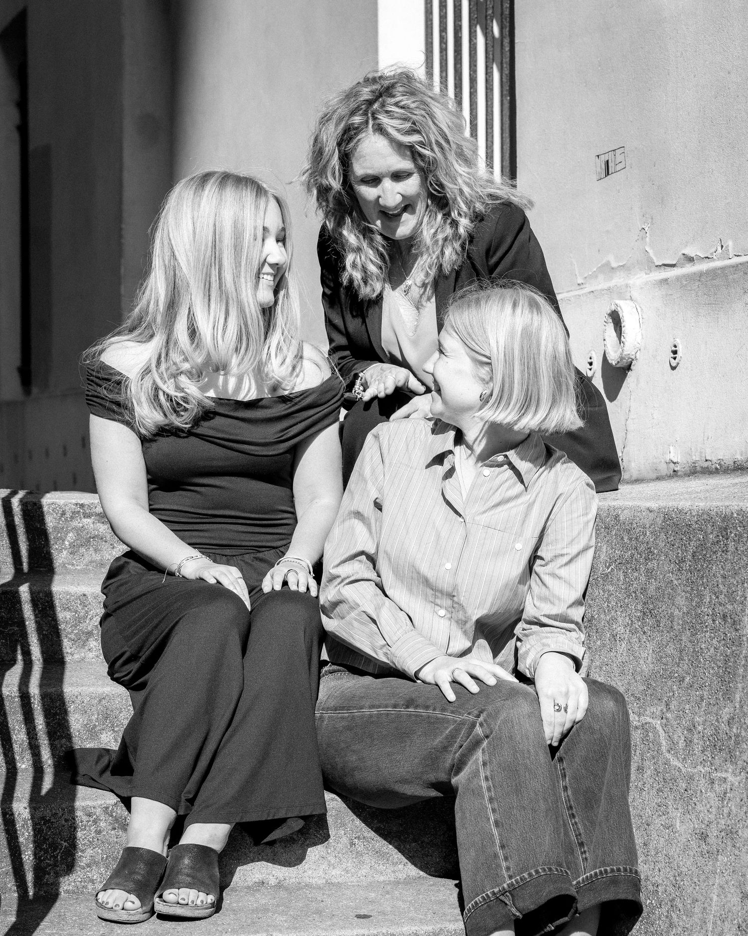 Black and white photo of three blonde women sitting on steps, laughing and sharing a joyful moment together outdoors.