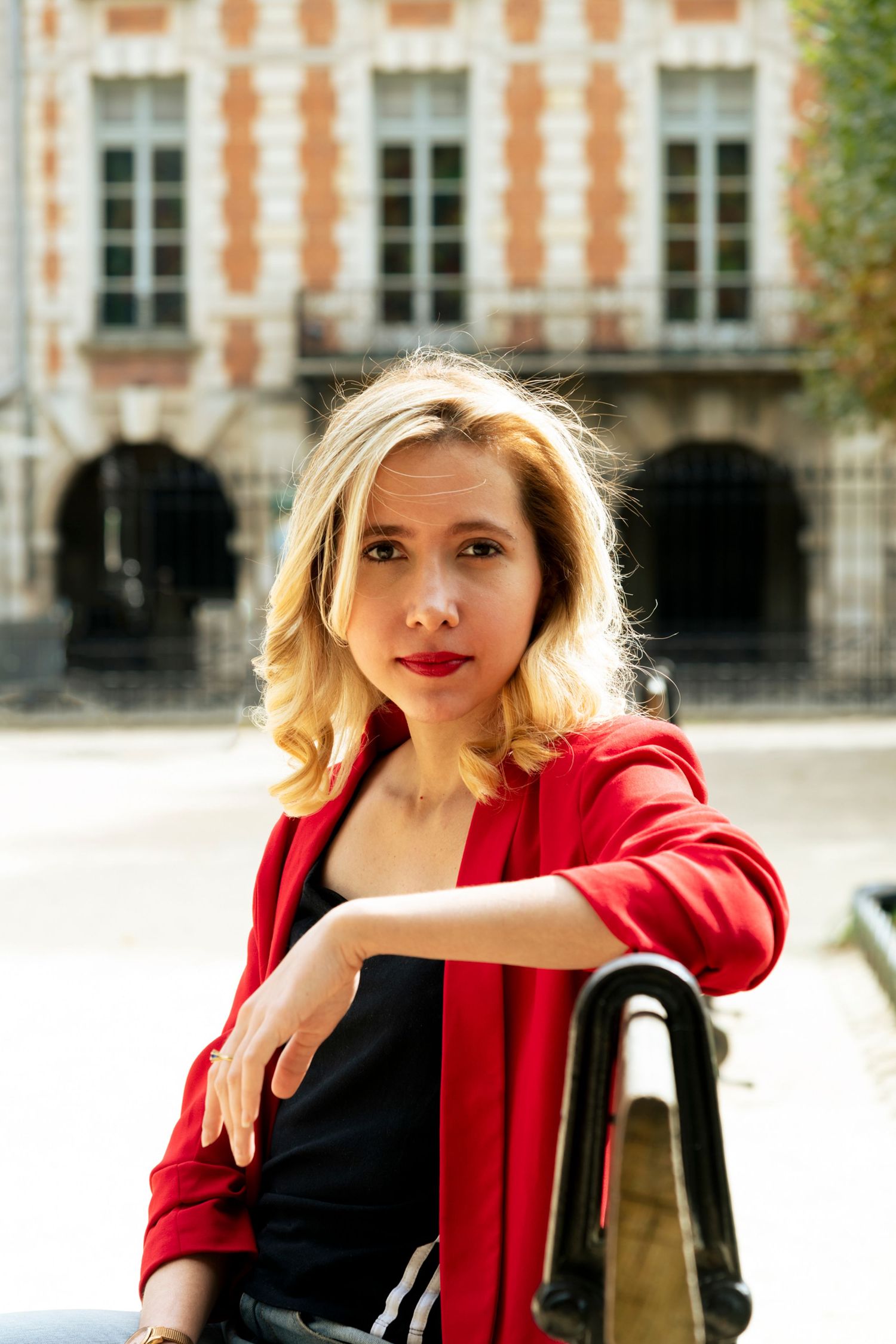 Blonde woman in red blazer sitting outdoors with historic European building facade in background.