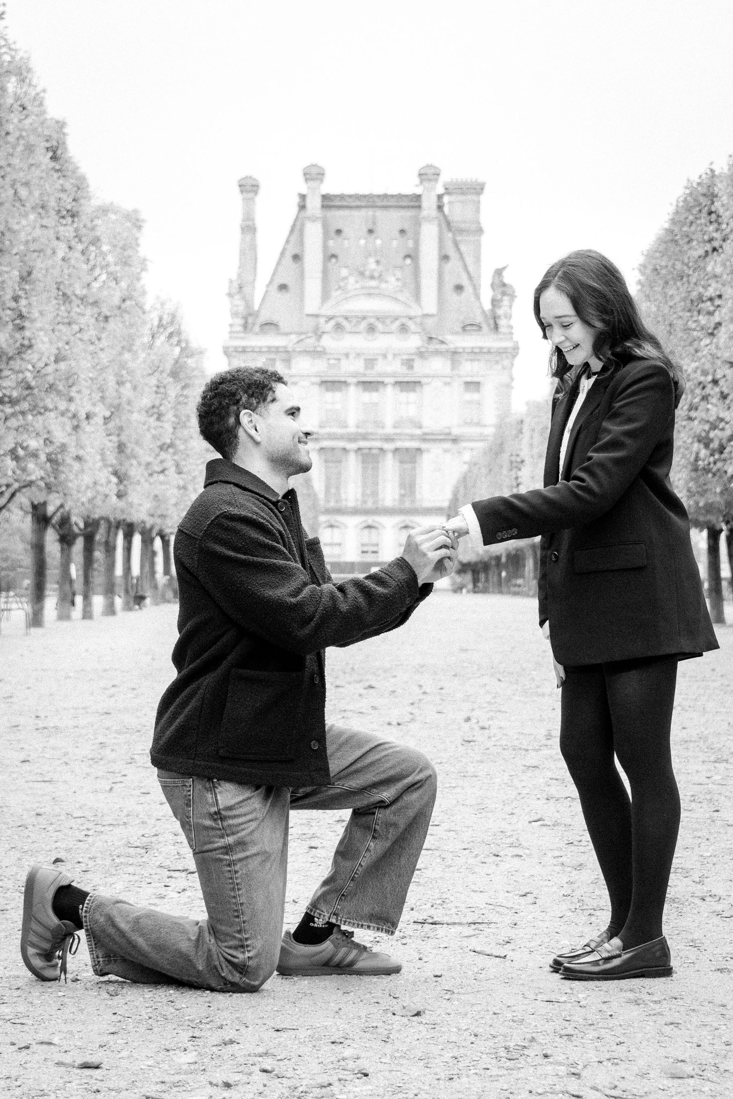 Man proposing on one knee to woman in Paris park with Louvre building backdrop, black and white photo.
