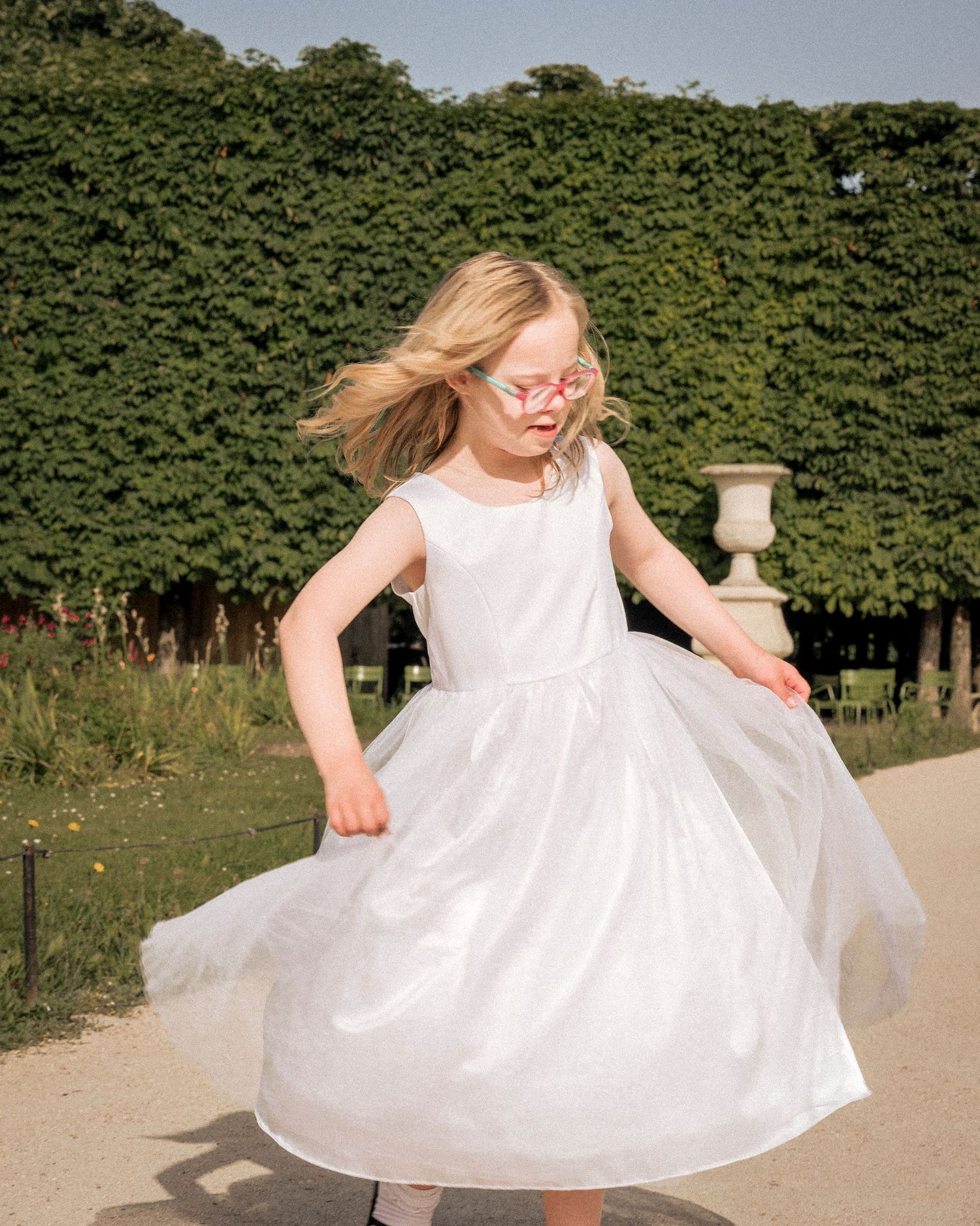 Young girl in white tulle dress twirling joyfully in formal garden wearing pink glasses.