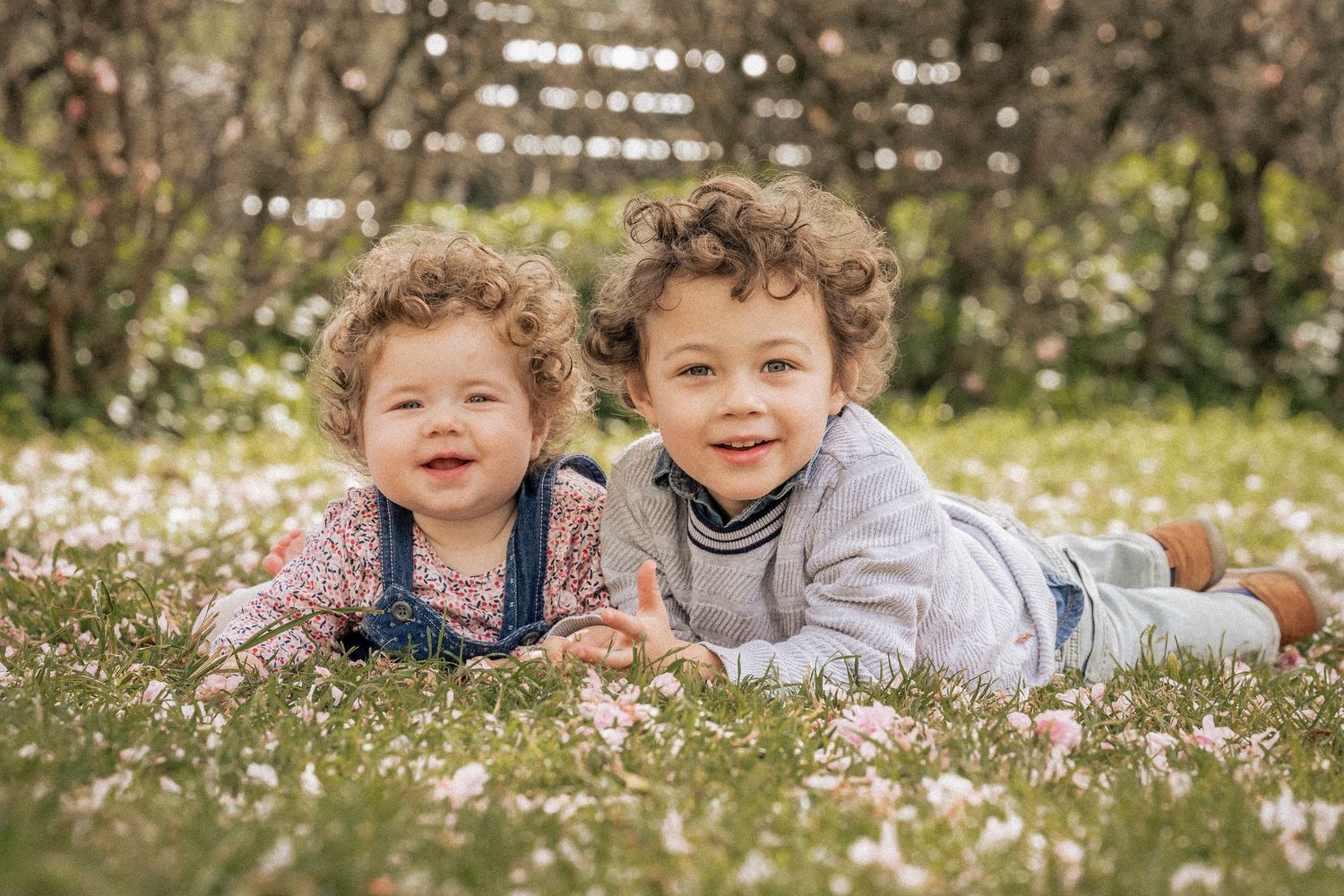 Two curly-haired toddlers lying in a flower-covered grass field, smiling at the camera in spring.