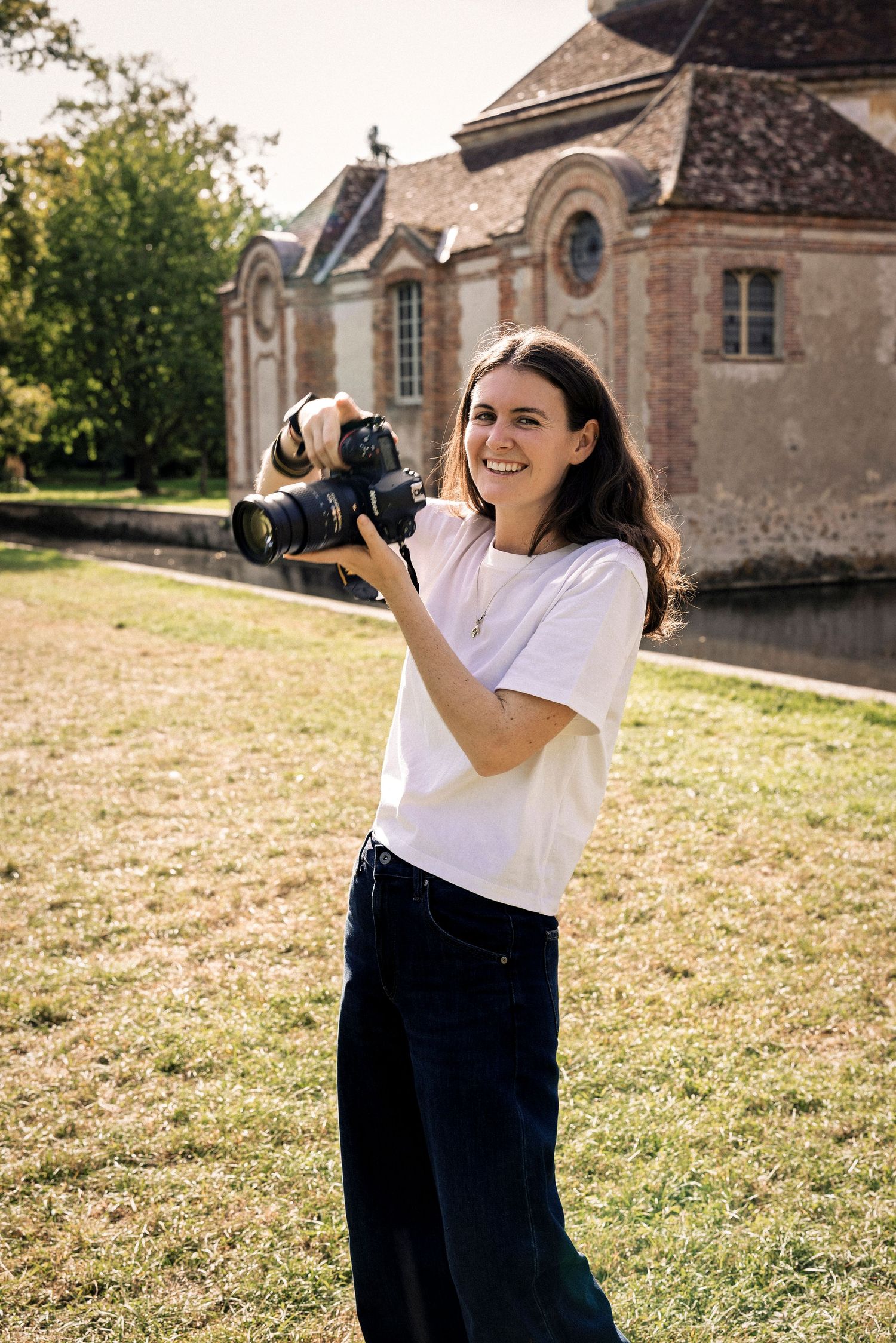 Happy young female photographer smiling while holding DSLR camera outdoors near historic stone building.
