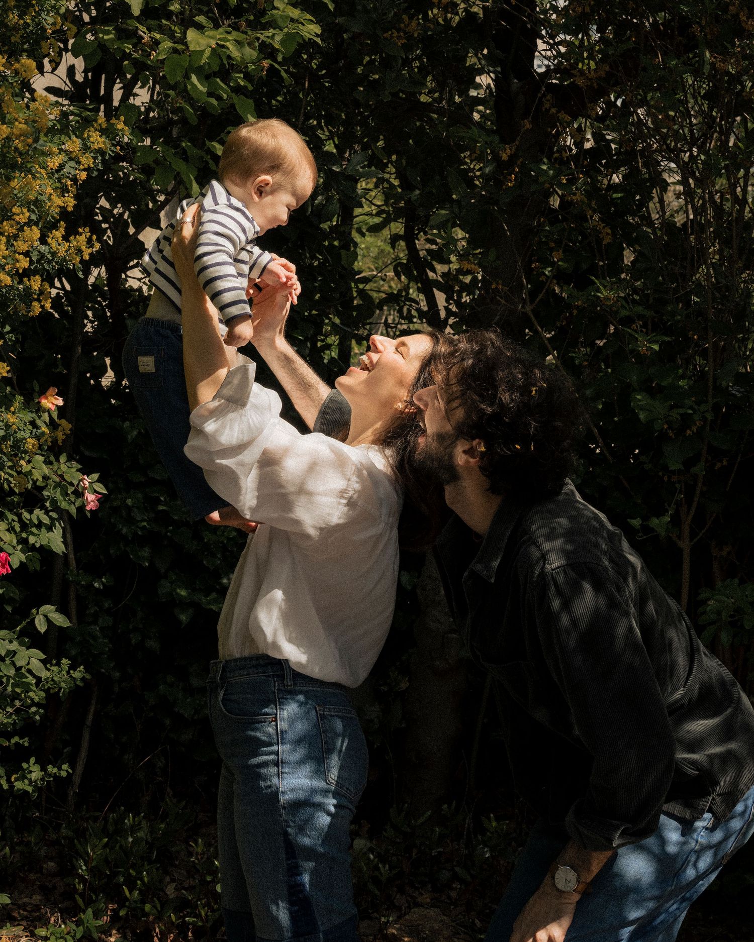 A couple joyfully lifts their baby in a sunlit garden surrounded by lush green trees.