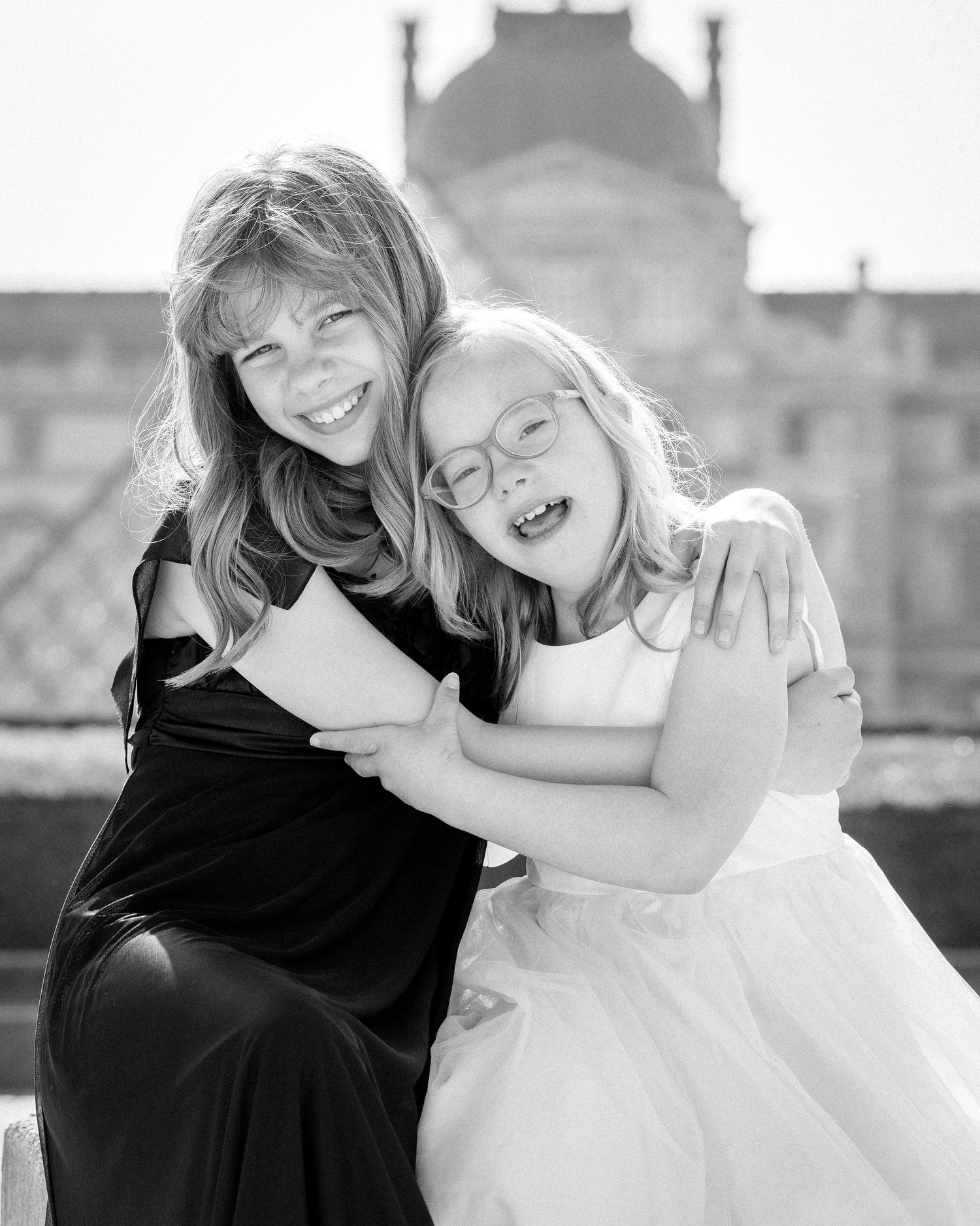 Two girls hugging and laughing in black and white photo with historic building in background.