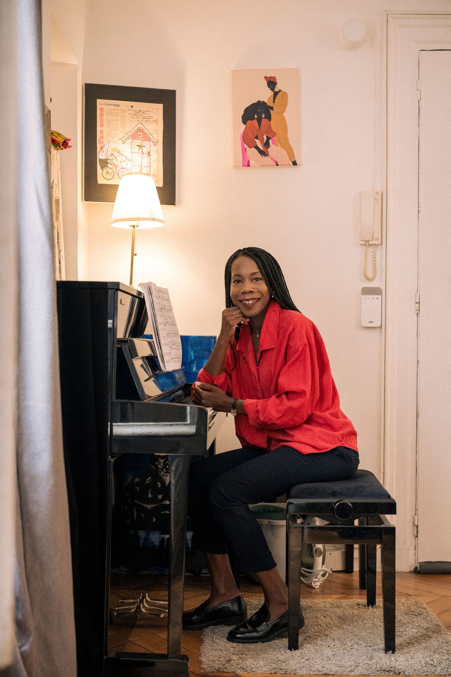 Smiling woman in a red blouse sitting at an upright piano in a cozy decorated room.