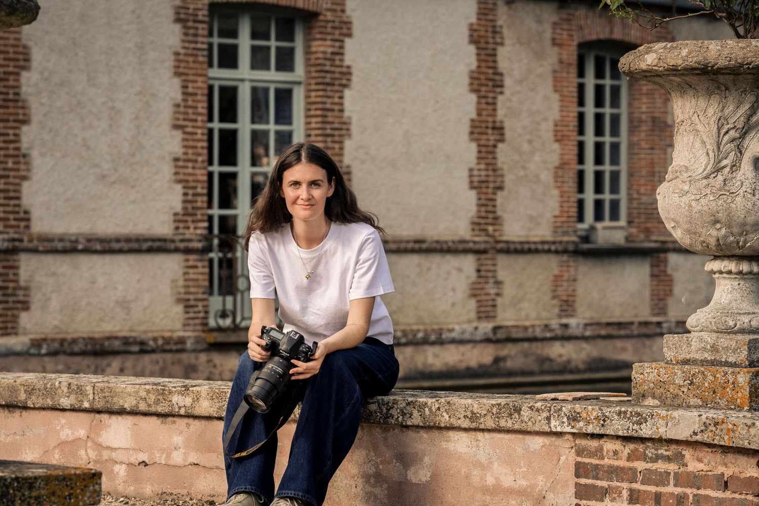 Young woman photographer sitting on stone wall holding camera outdoors near historic brick building.