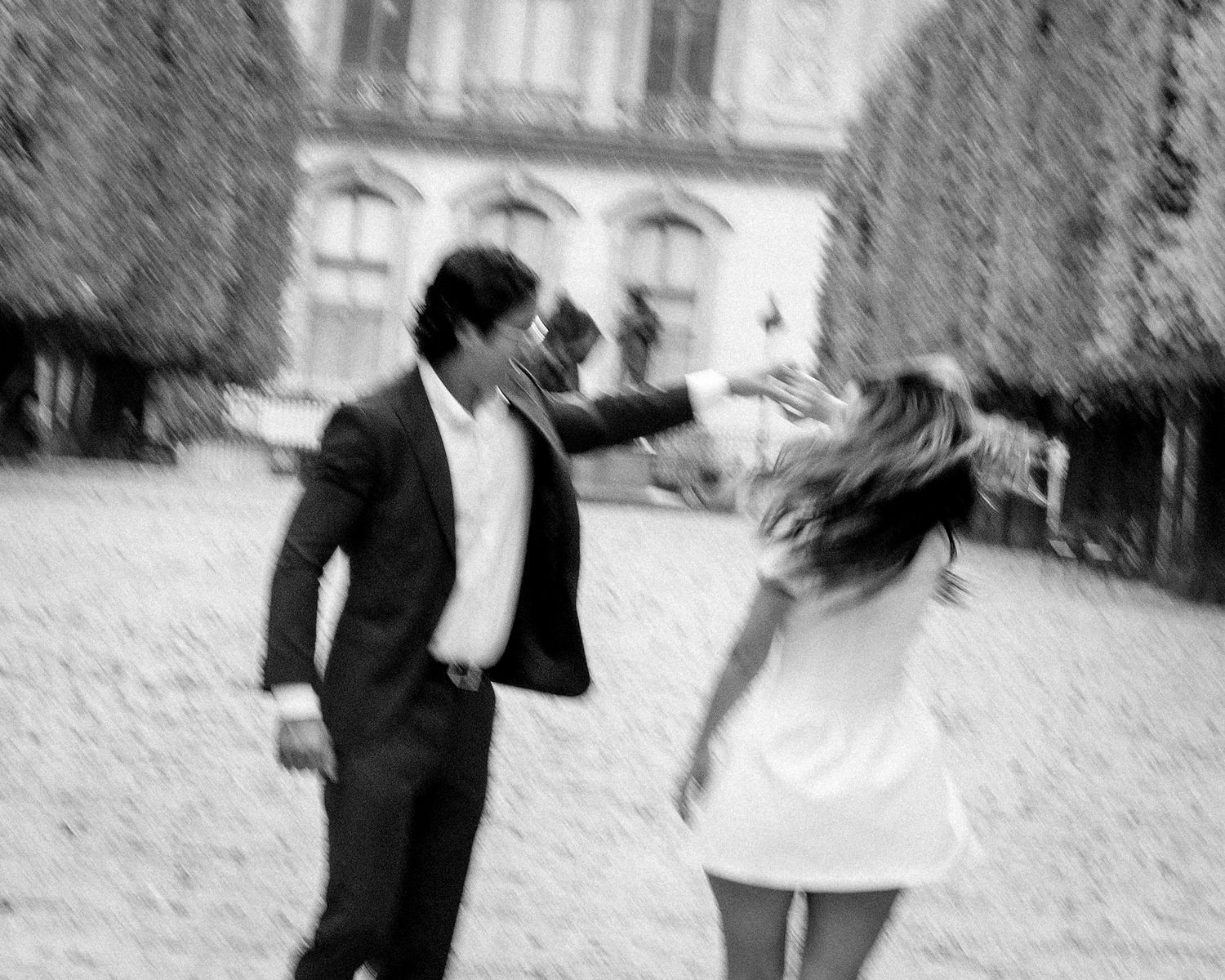 Blurry black and white photo of couple dancing playfully in courtyard.