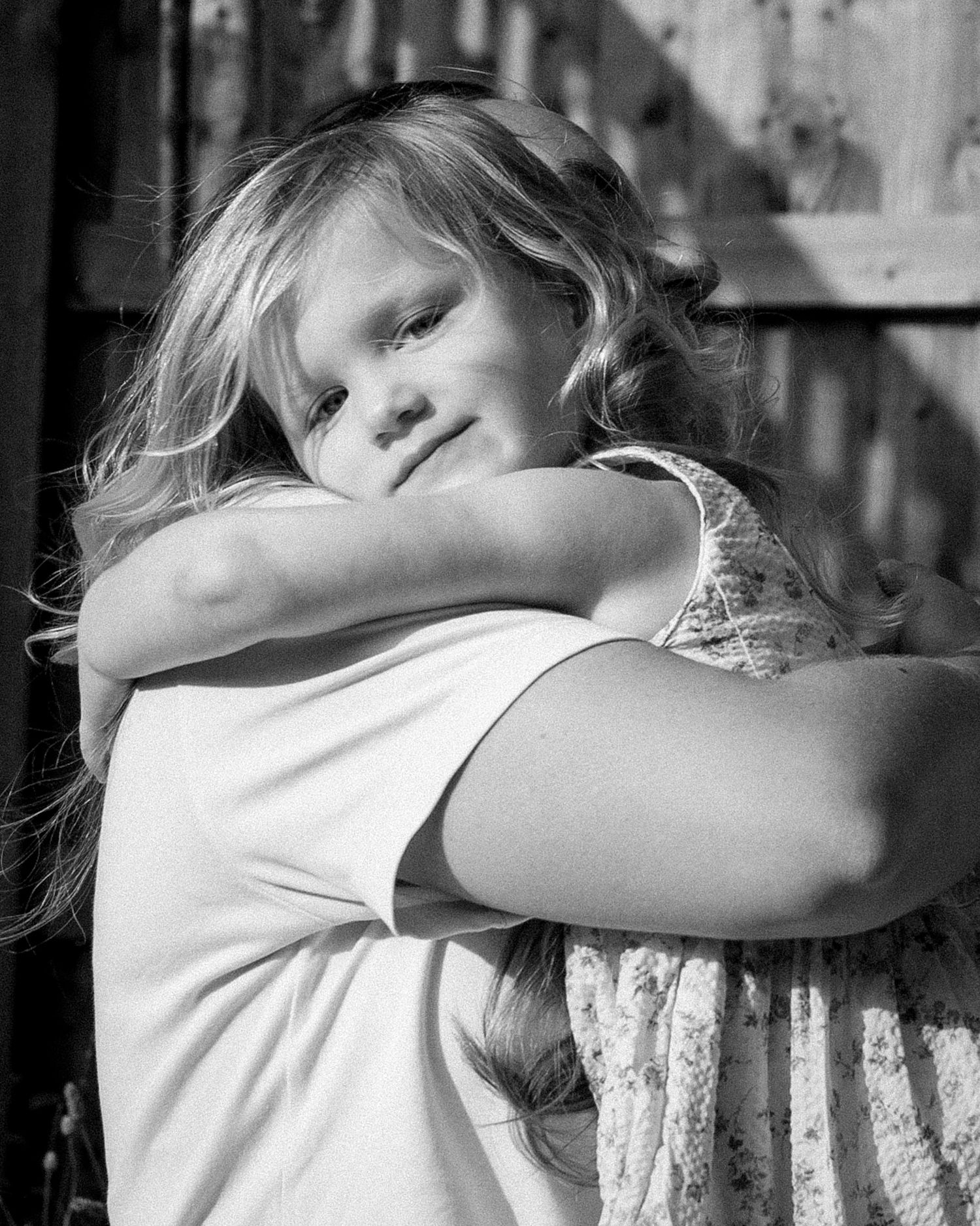Black and white portrait of a young blonde girl hugging someone tightly, smiling softly in warm sunlight outdoors.