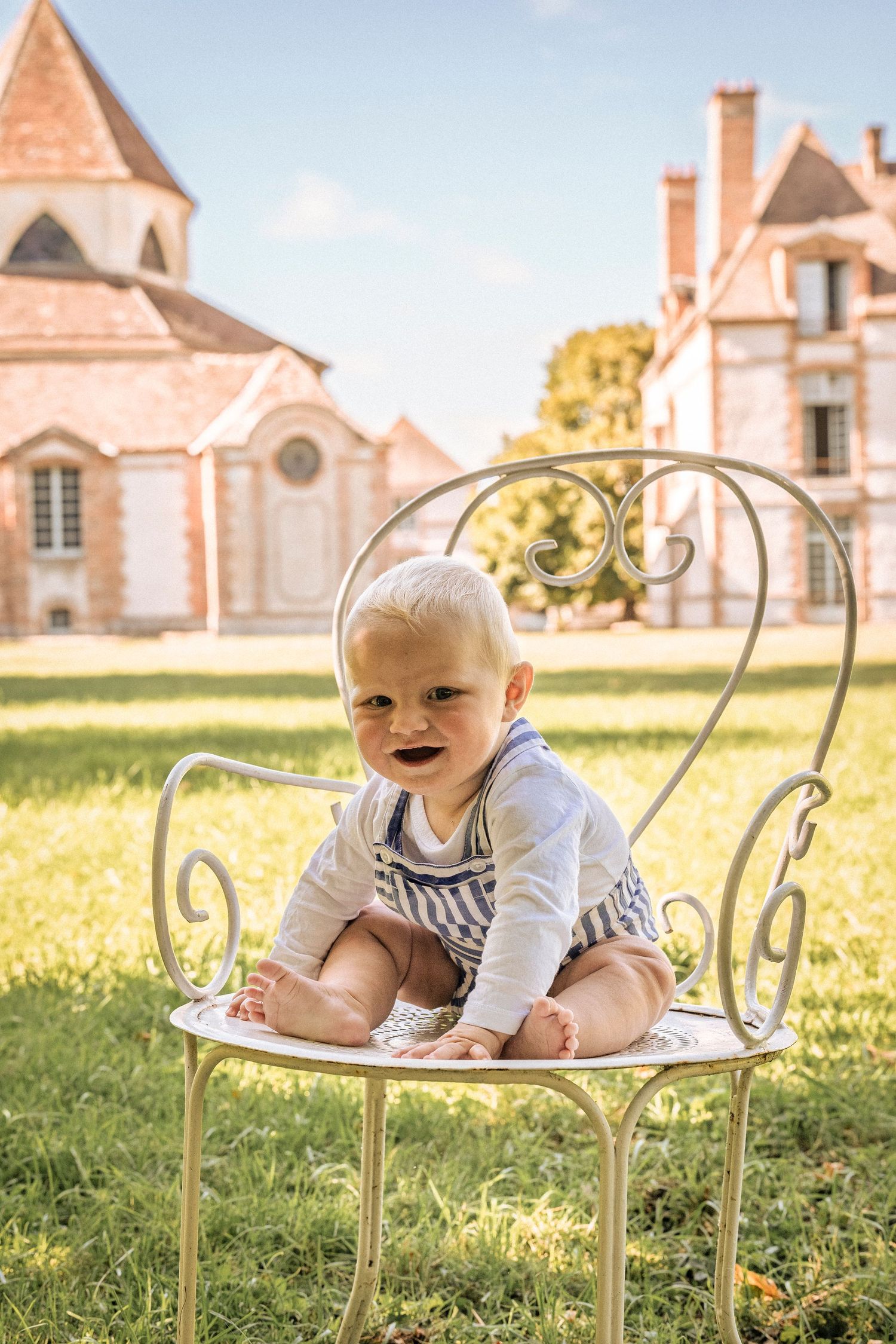 Adorable blonde baby sitting on a white ornate metal chair on green grass in front of a historic French château.