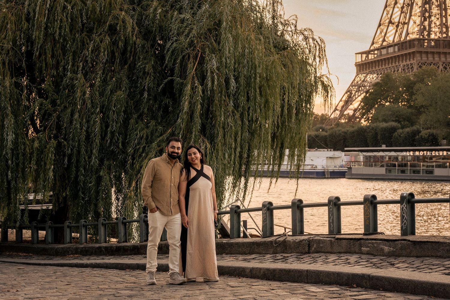 Happy couple embraces under a weeping willow tree near the Seine River with the Eiffel Tower at sunset.