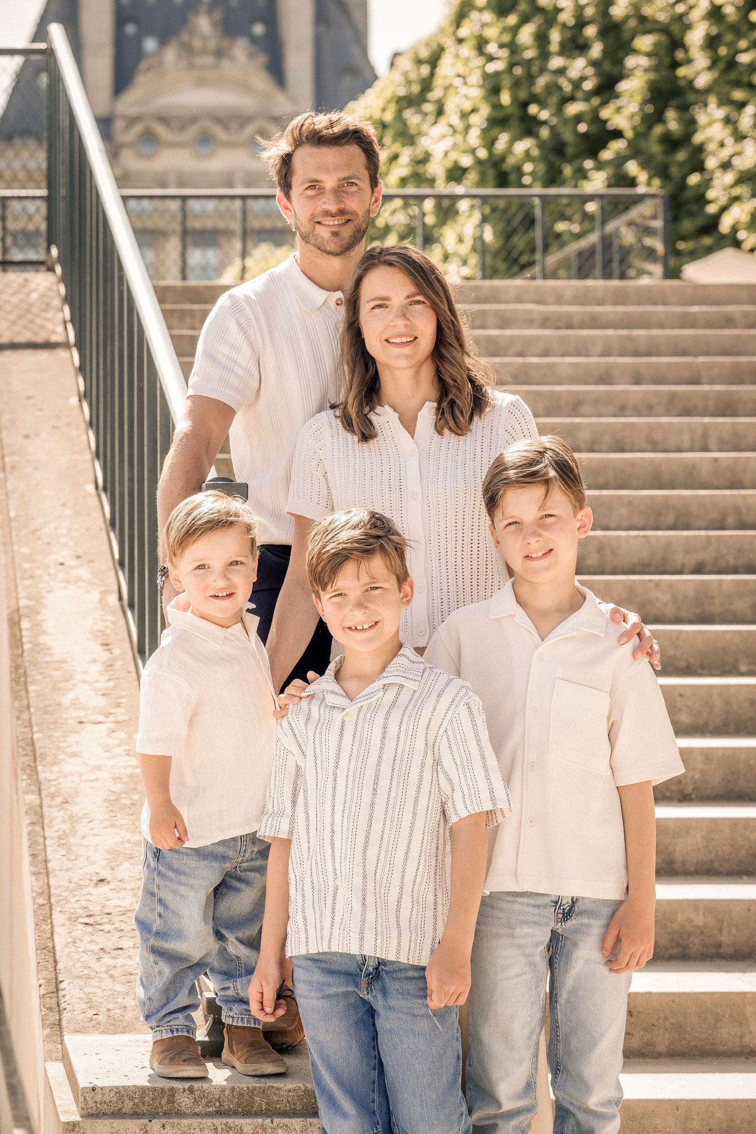 Family of five in white outfits posing on sunny stone steps near a Paris landmark.