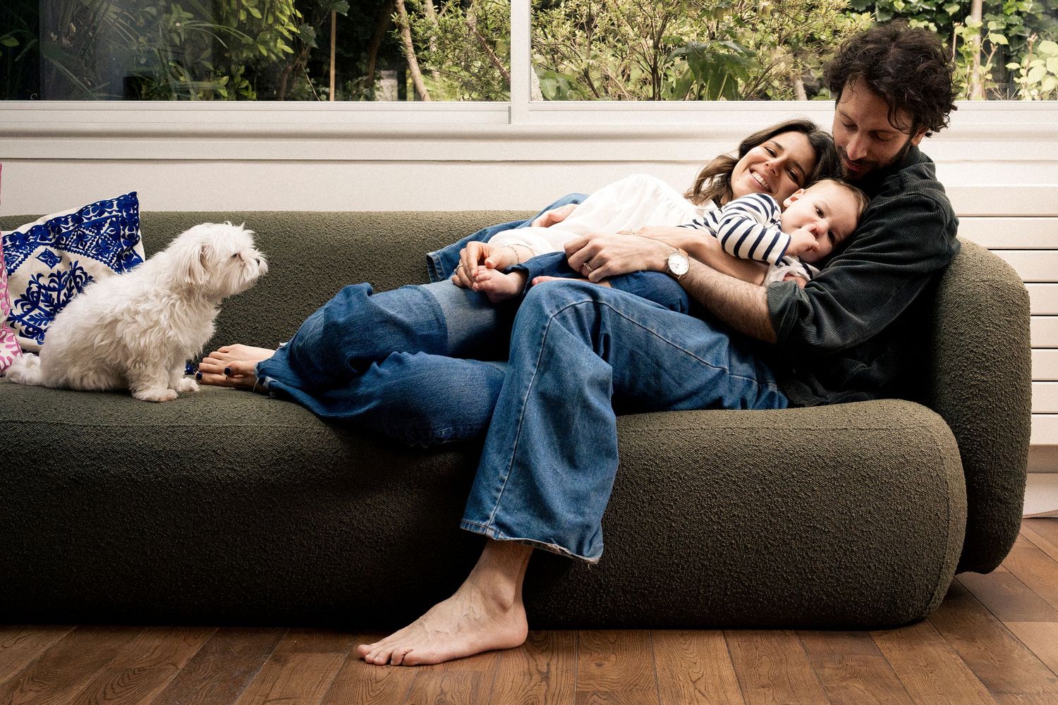 Family relaxing together on a cozy green sofa with a white fluffy dog nearby on hardwood floors.