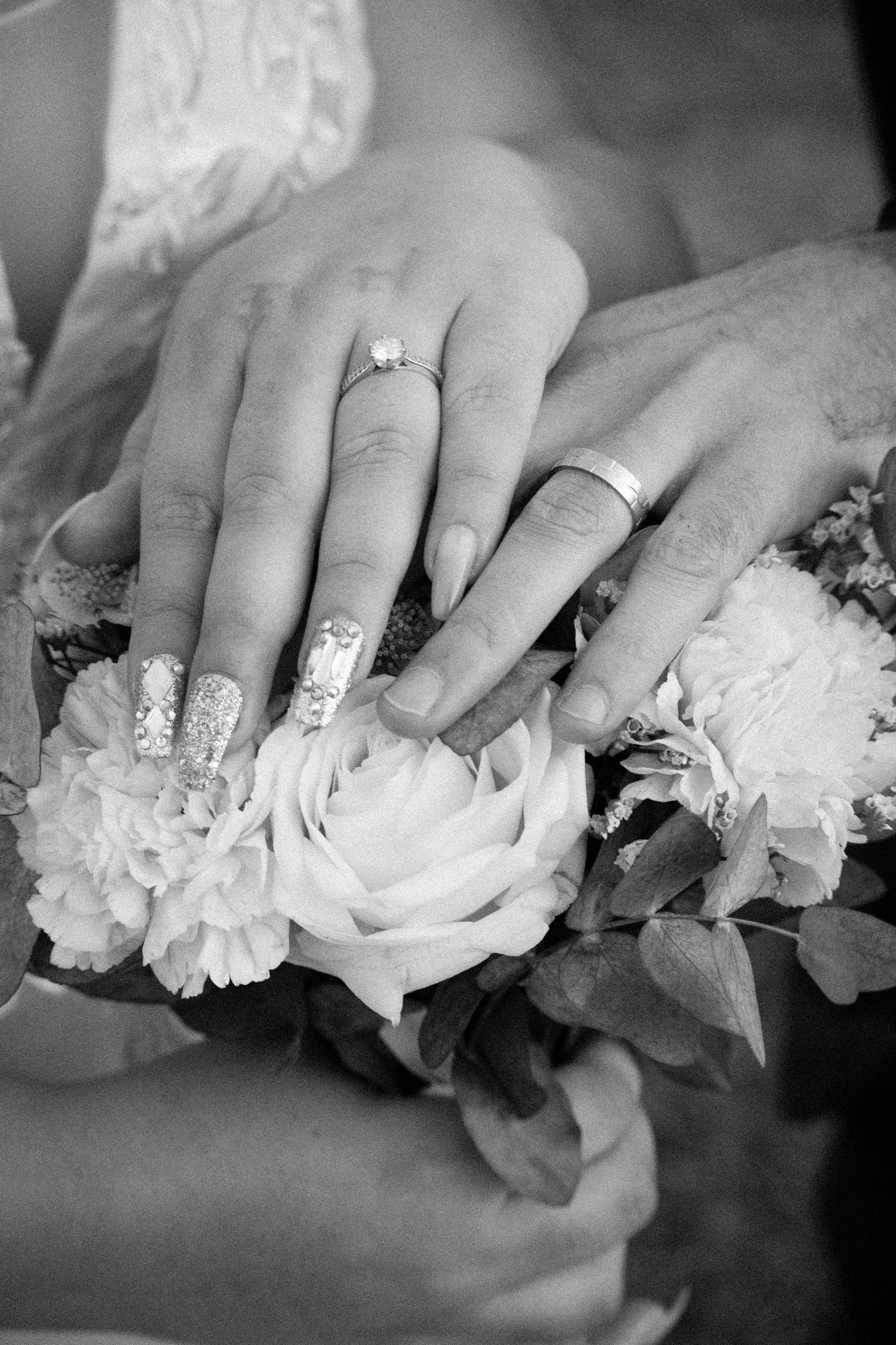 Black and white close-up of newlyweds hands with wedding rings resting on bridal bouquet.