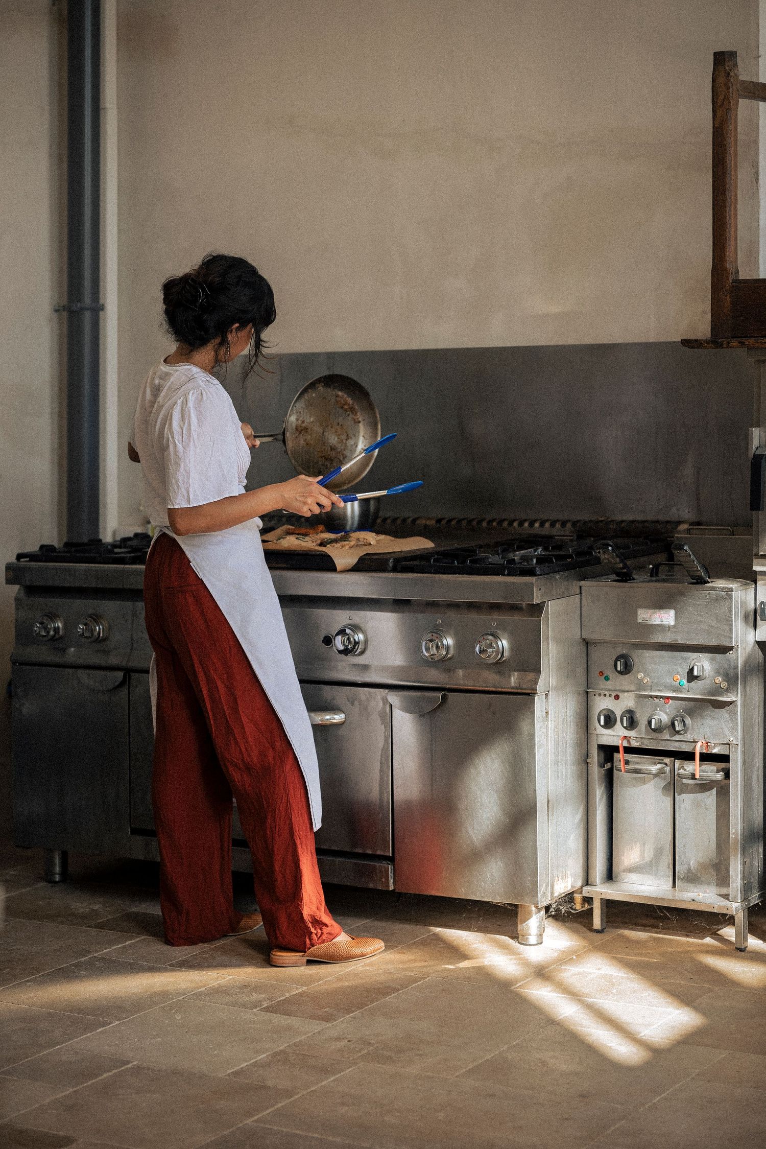 Chef in white apron cooking in a professional stainless steel kitchen with natural sunlight.