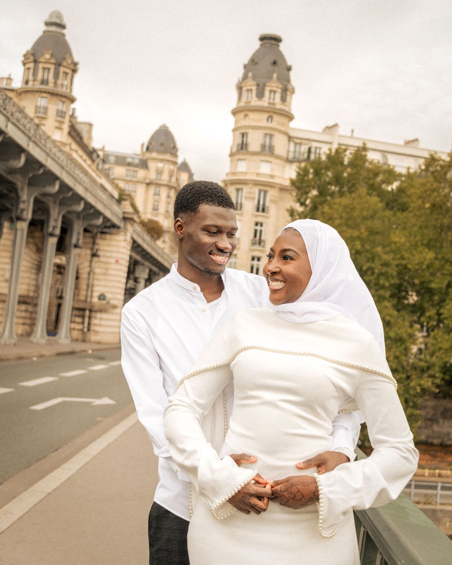 A couple dressed in white stands on a Paris bridge, smiling together with a grand building behind them.