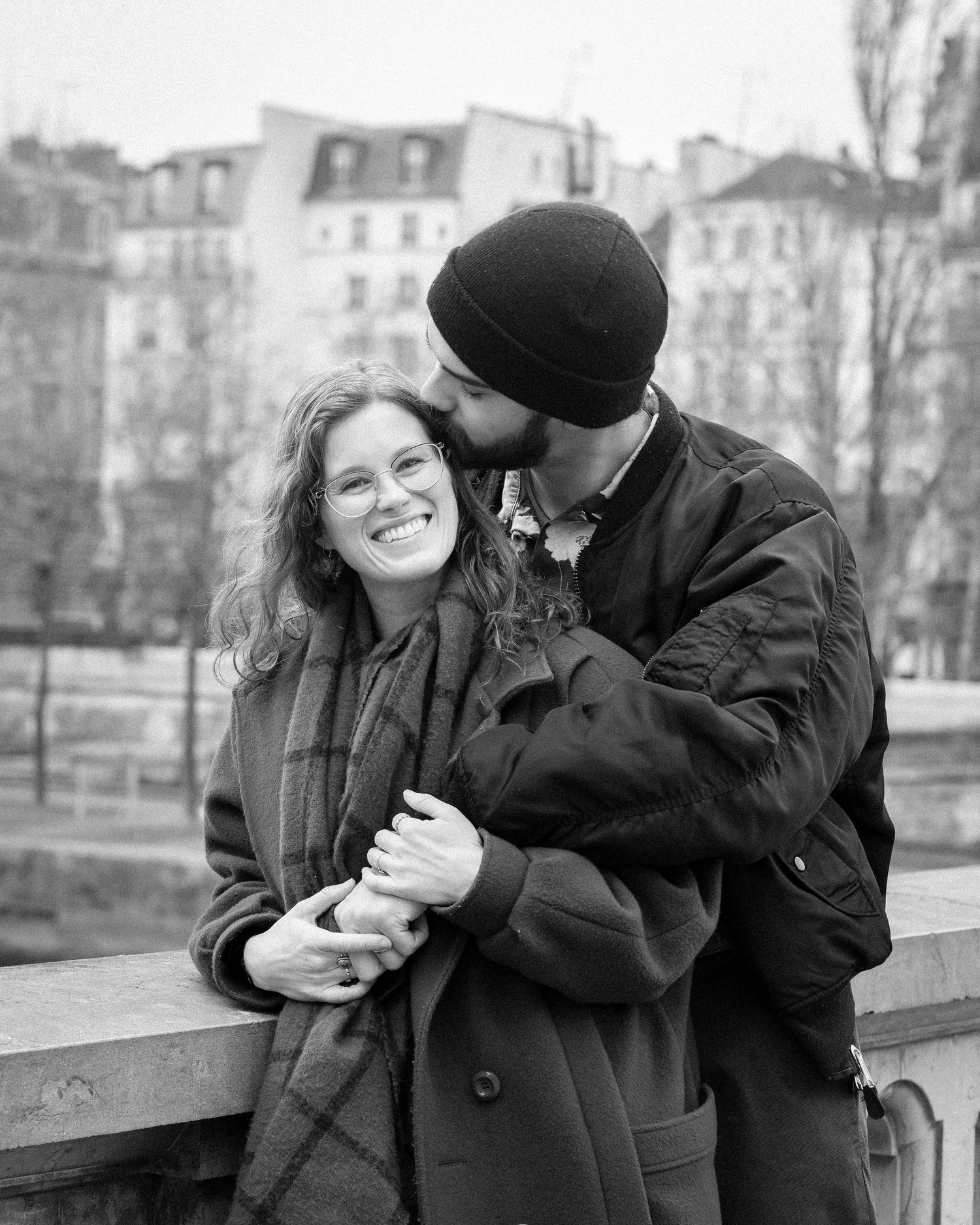 Black and white photo of a couple embracing on a Paris bridge, man kissing woman's cheek as she smiles.