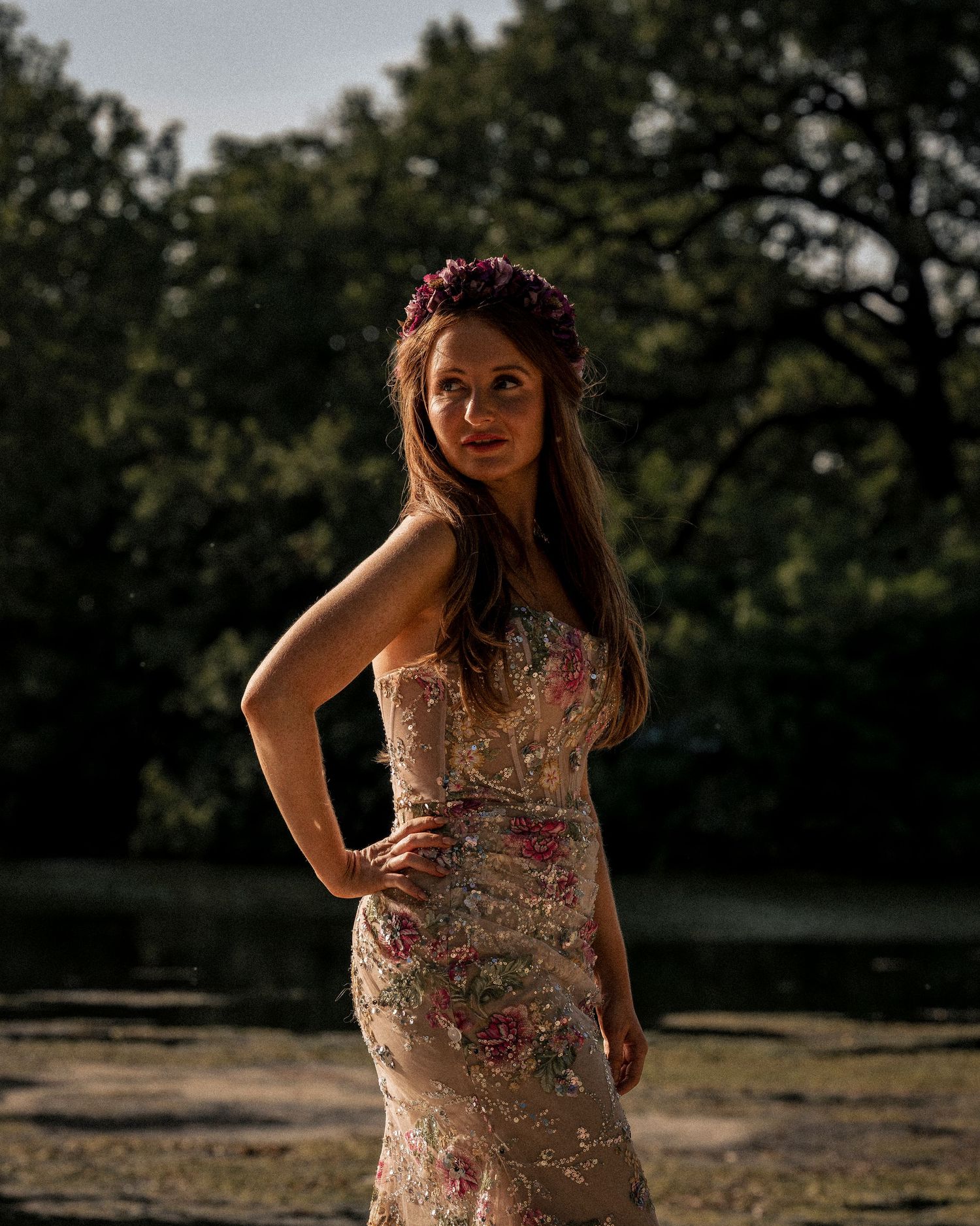 Woman in floral embroidered dress with flower crown posing in a shaded garden at sunset.