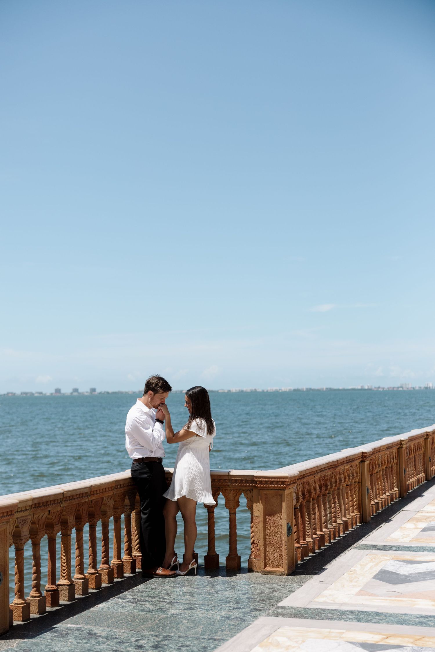 Couple holding hands on a waterfront terrace with ornate stone balustrade and blue ocean backdrop.