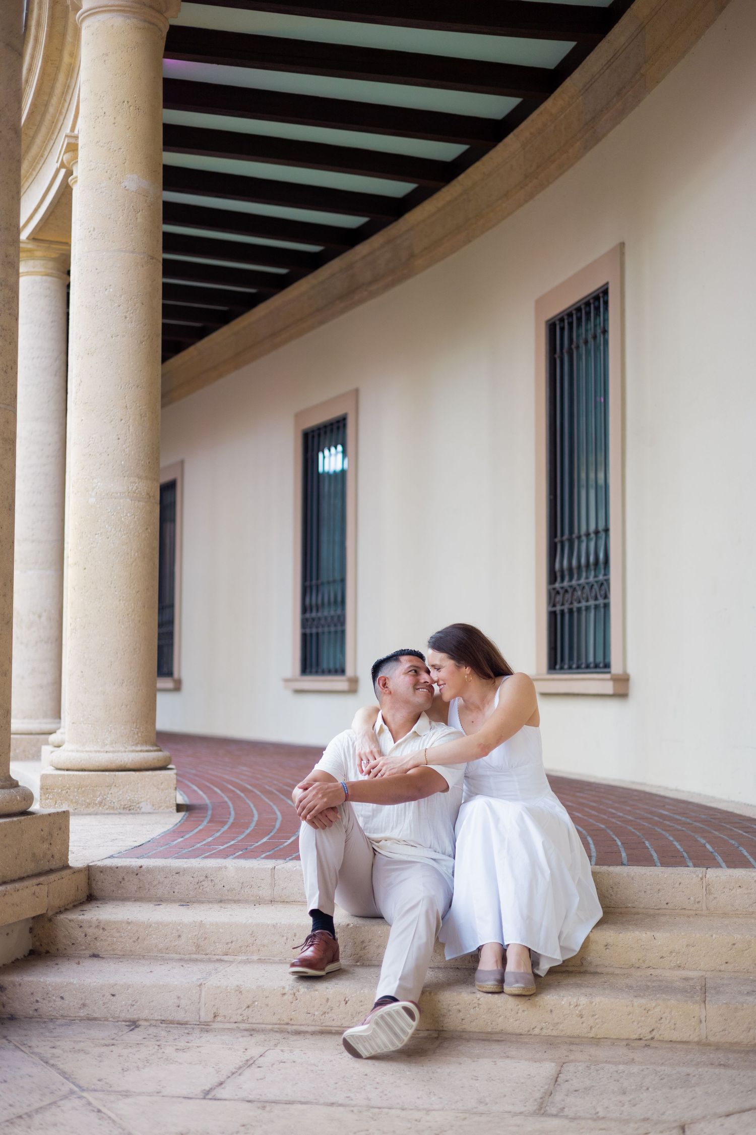 Couple sharing a tender kiss while sitting on steps between classic columns at a Mediterranean-style building.