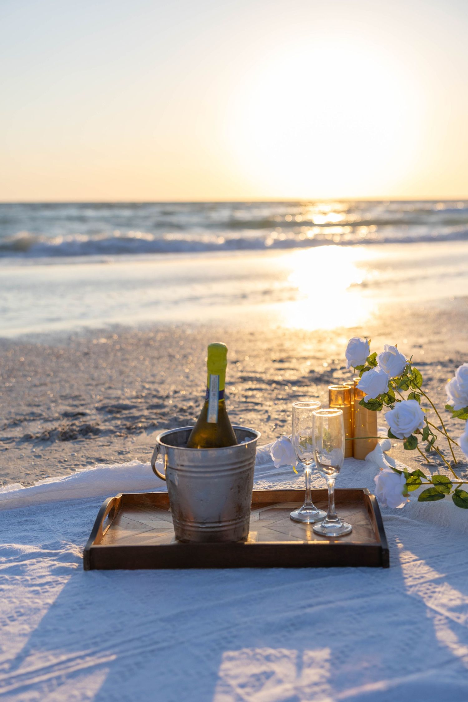 A champagne bottle in ice bucket with two glasses and white roses on a tray set on a beach at golden sunset.