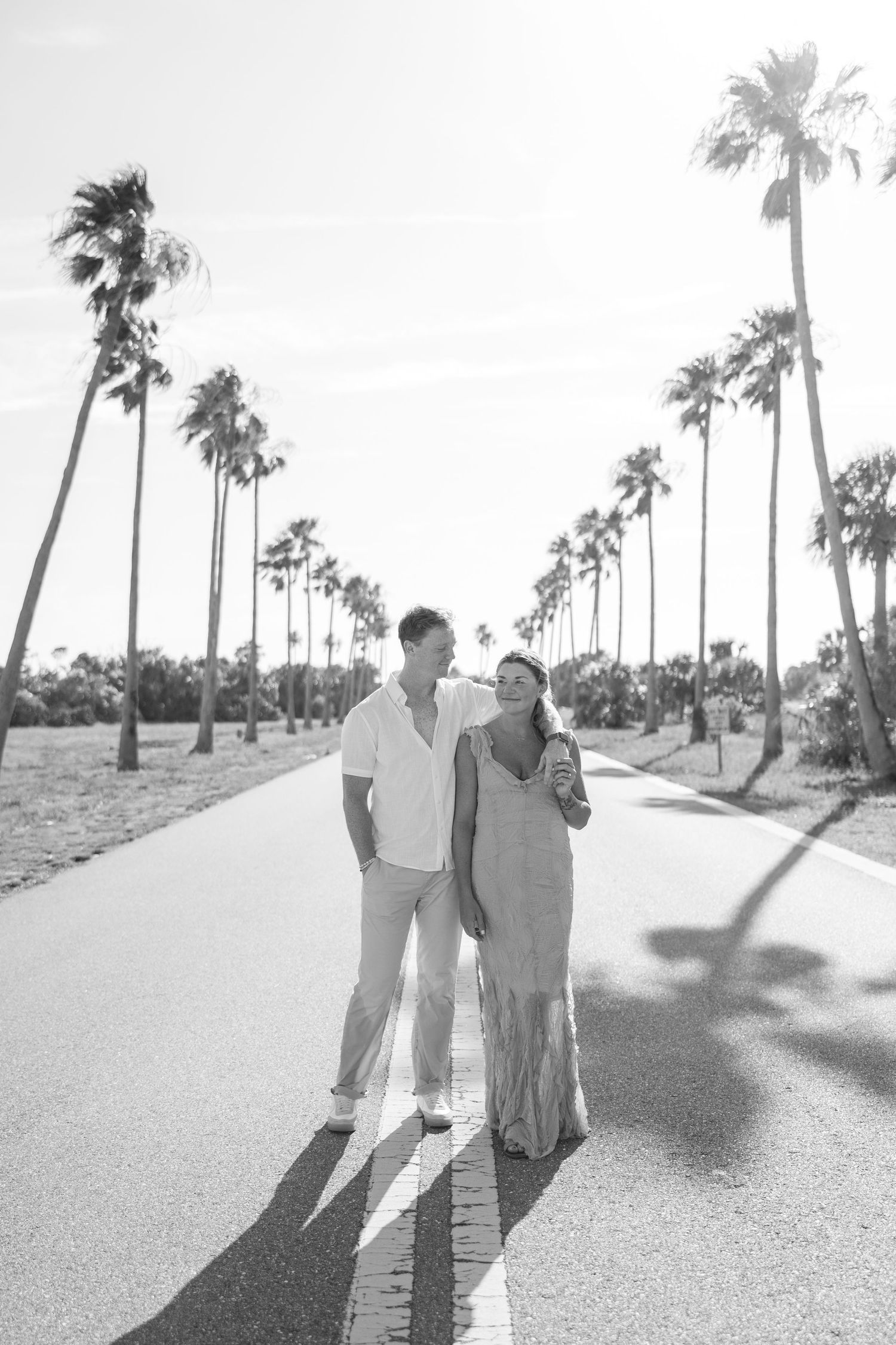 Black and white photo of couple walking down a palm tree-lined road in sunshine.