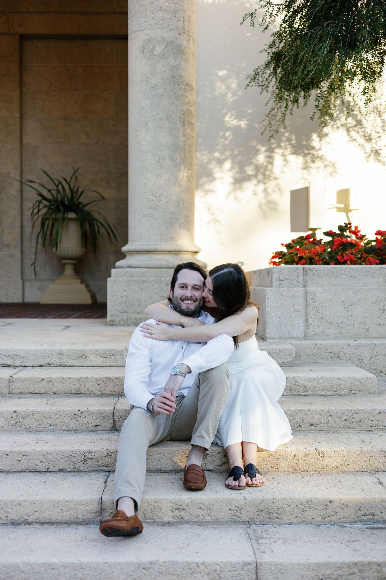 Couple sitting on marble steps embracing, woman kissing man's cheek near stone columns and red flowers.