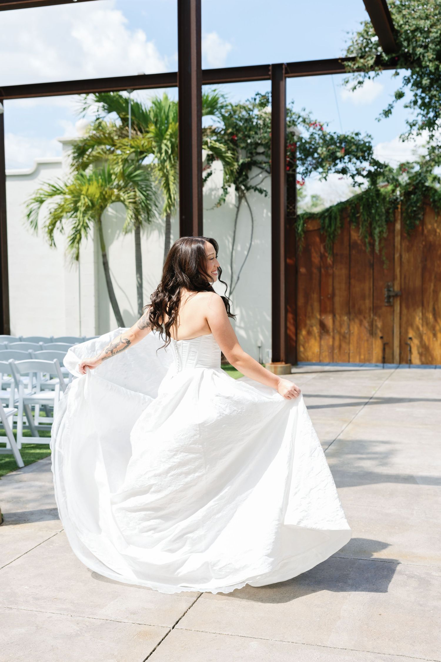 Bride in a flowing white gown twirls joyfully on a sunlit patio with tropical trees in the background.