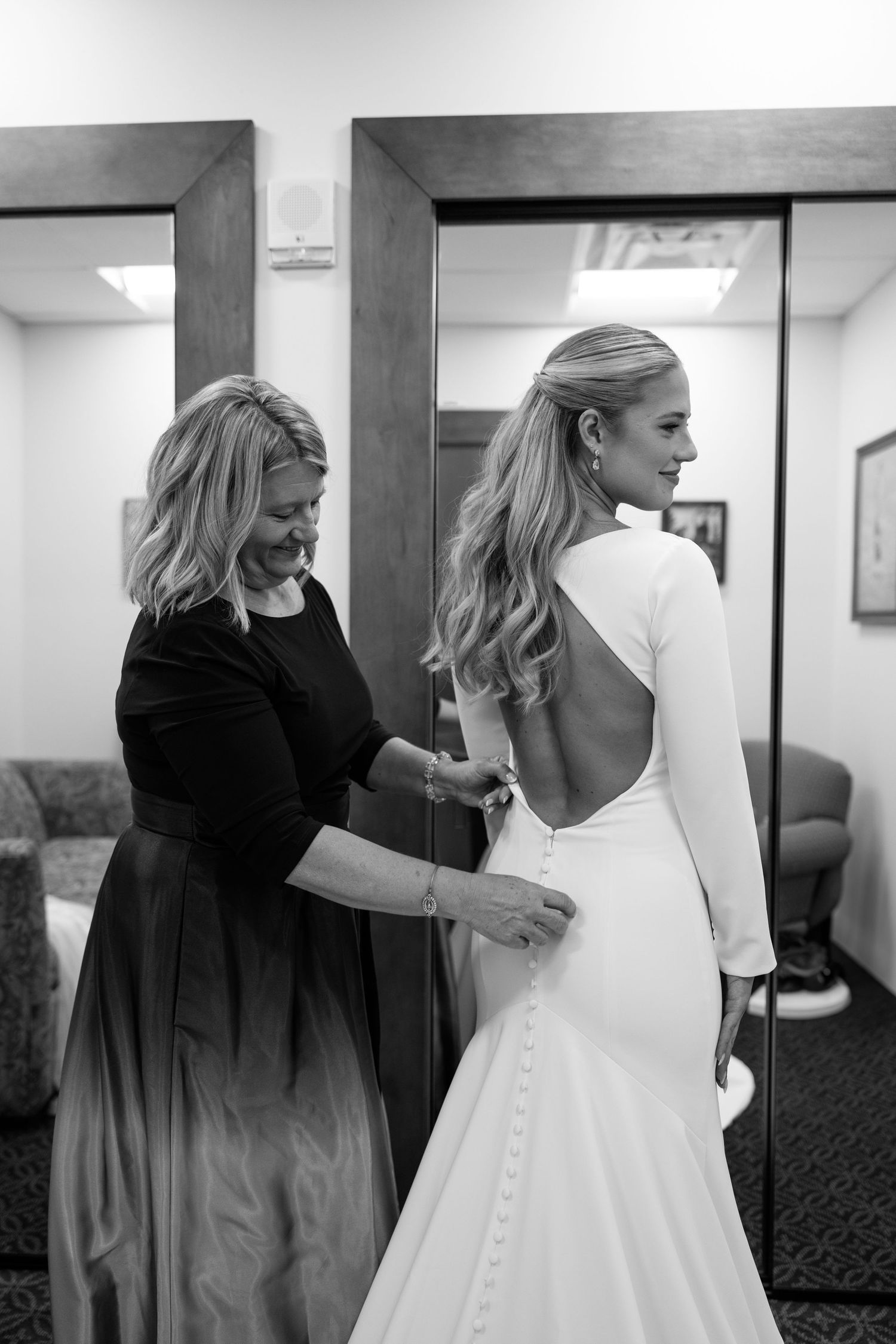 Black and white photo of mother helping bride button up elegant open-back wedding gown.