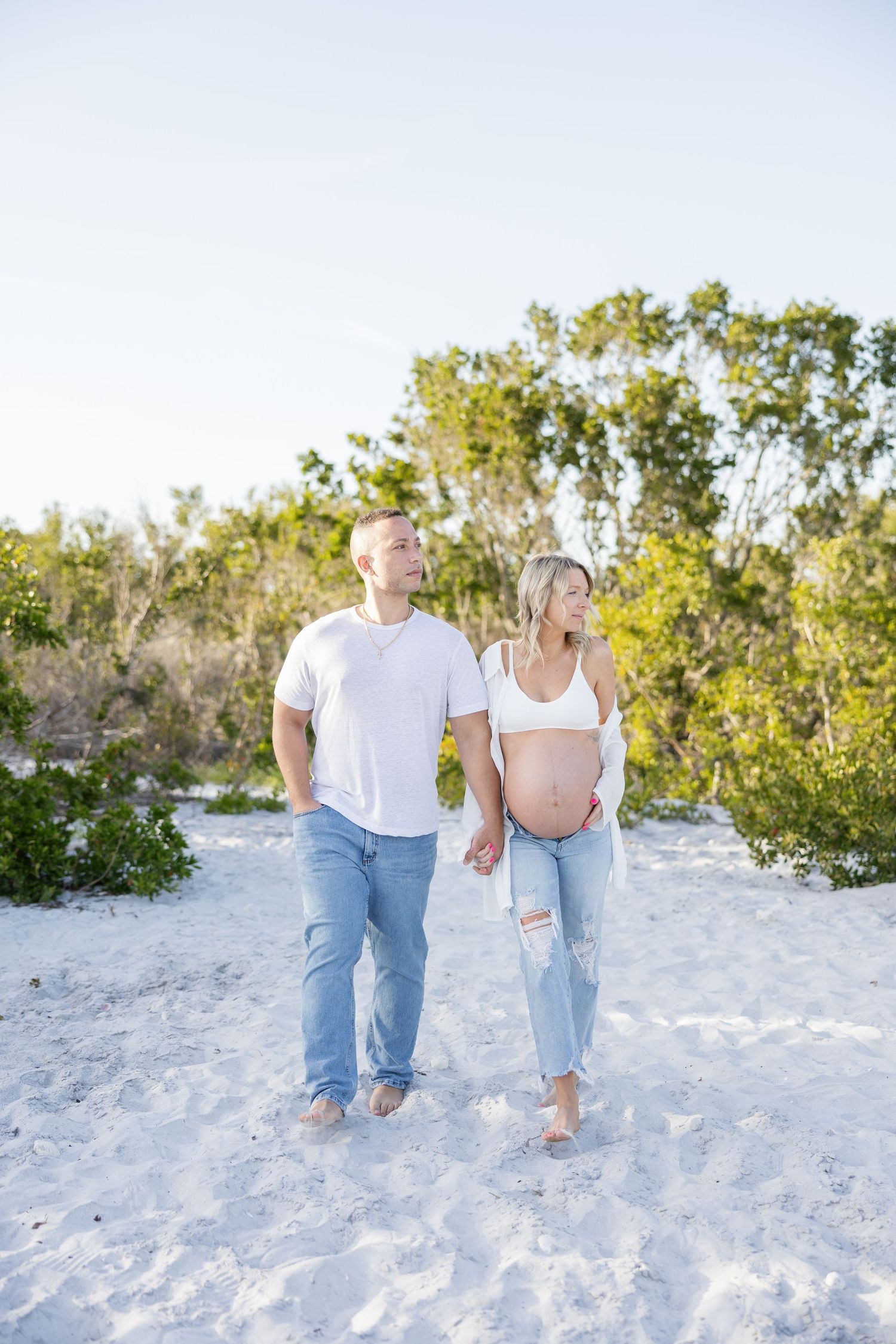 Pregnant couple holding hands walking on white sand beach surrounded by lush green trees.