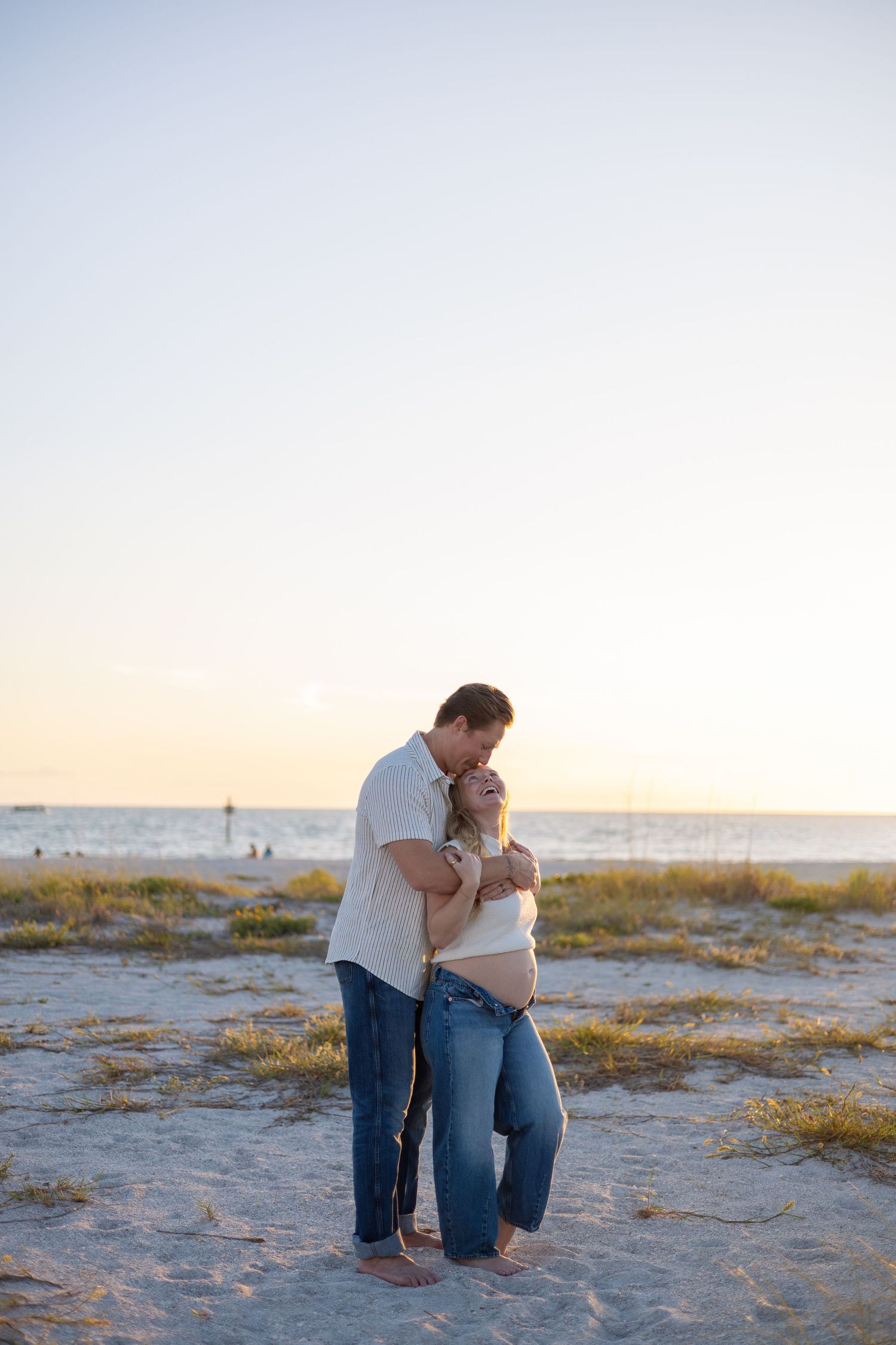 Couple embracing at golden hour on sandy beach, pregnant woman leaning into partners kiss.