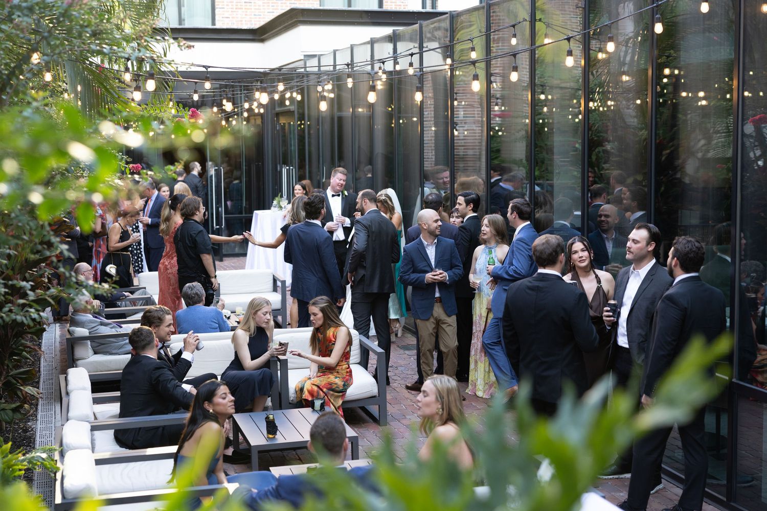 Elegantly dressed wedding guests mingle at outdoor courtyard cocktail hour with string lights and greenery.