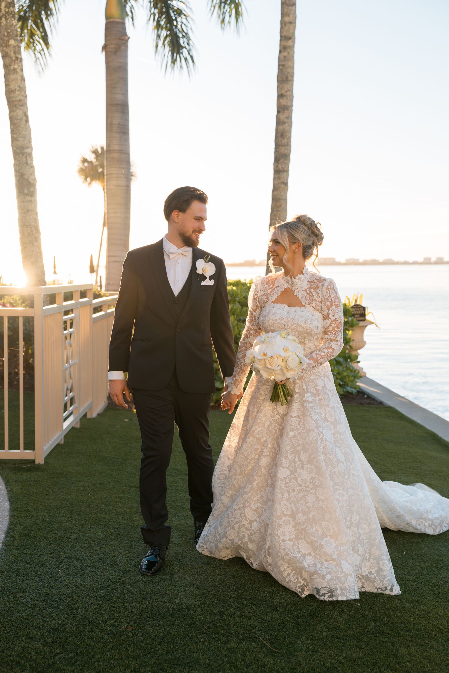 Bride in lace gown and groom in black tuxedo walk hand-in-hand at sunset near waterfront with palm trees.