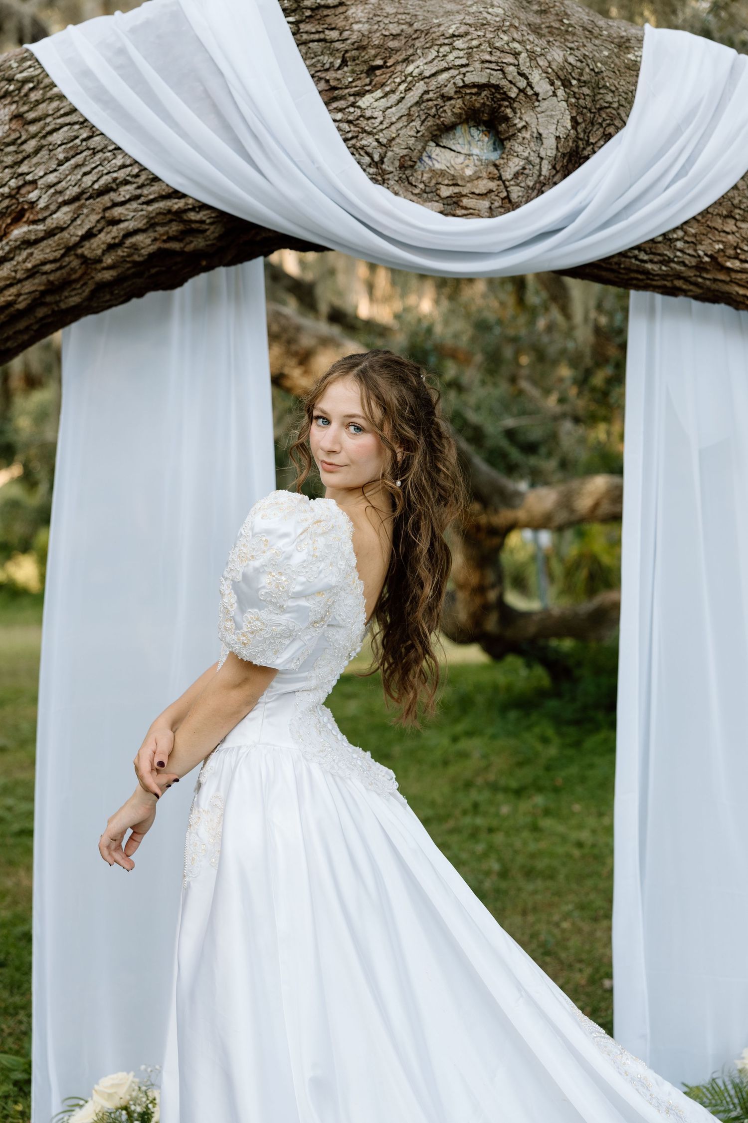 Bride in white lace puff-sleeve gown posing under draped white fabric and oak tree outdoors.