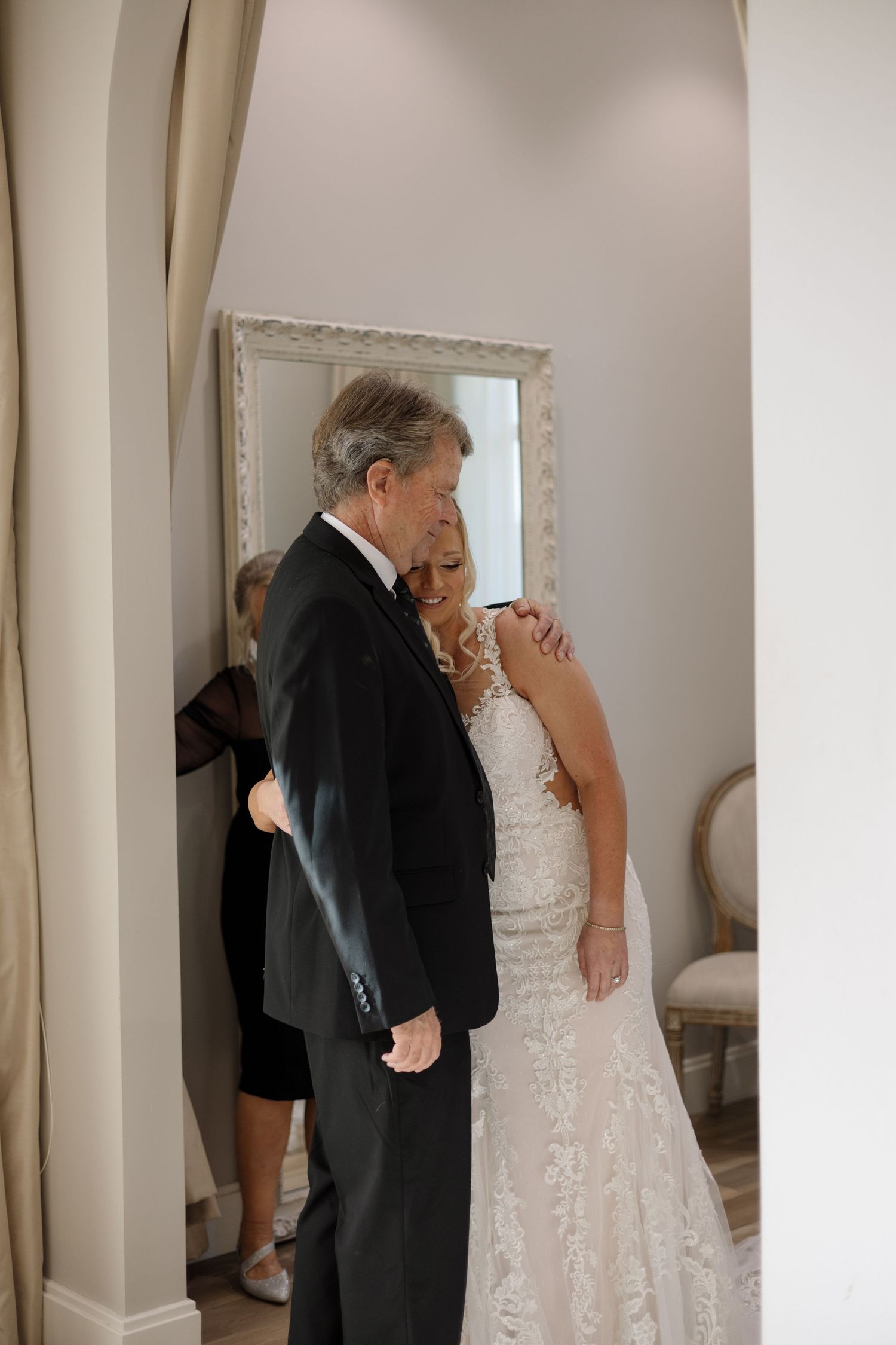 Emotional father embraces daughter in lace wedding gown during touching first look moment indoors.