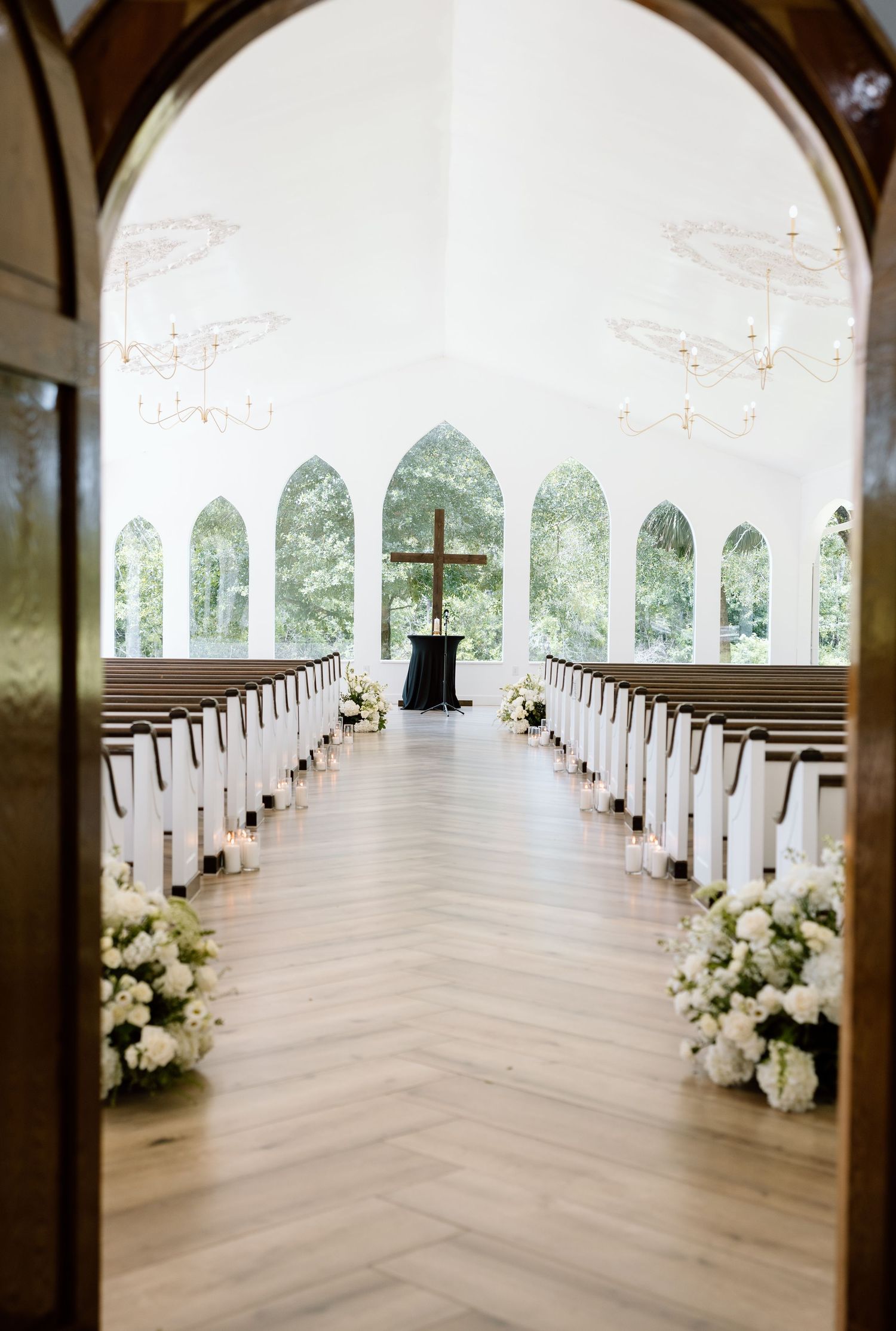 Elegant white chapel interior with wooden pews, herringbone floors, white floral arrangements, and arched windows.