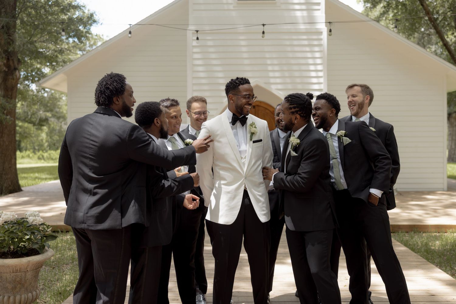 Groomsmen in black suits celebrate with groom in white tuxedo outside rustic white chapel.