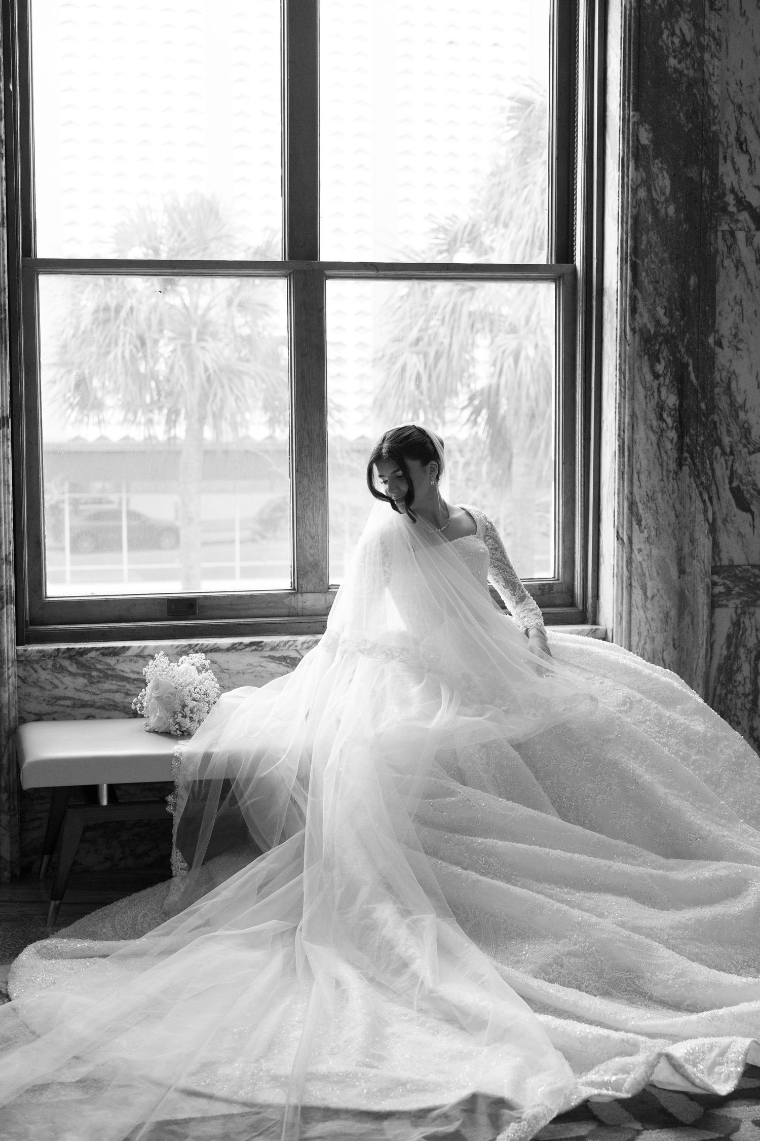 Black and white portrait of bride in flowing gown and veil seated by large window.