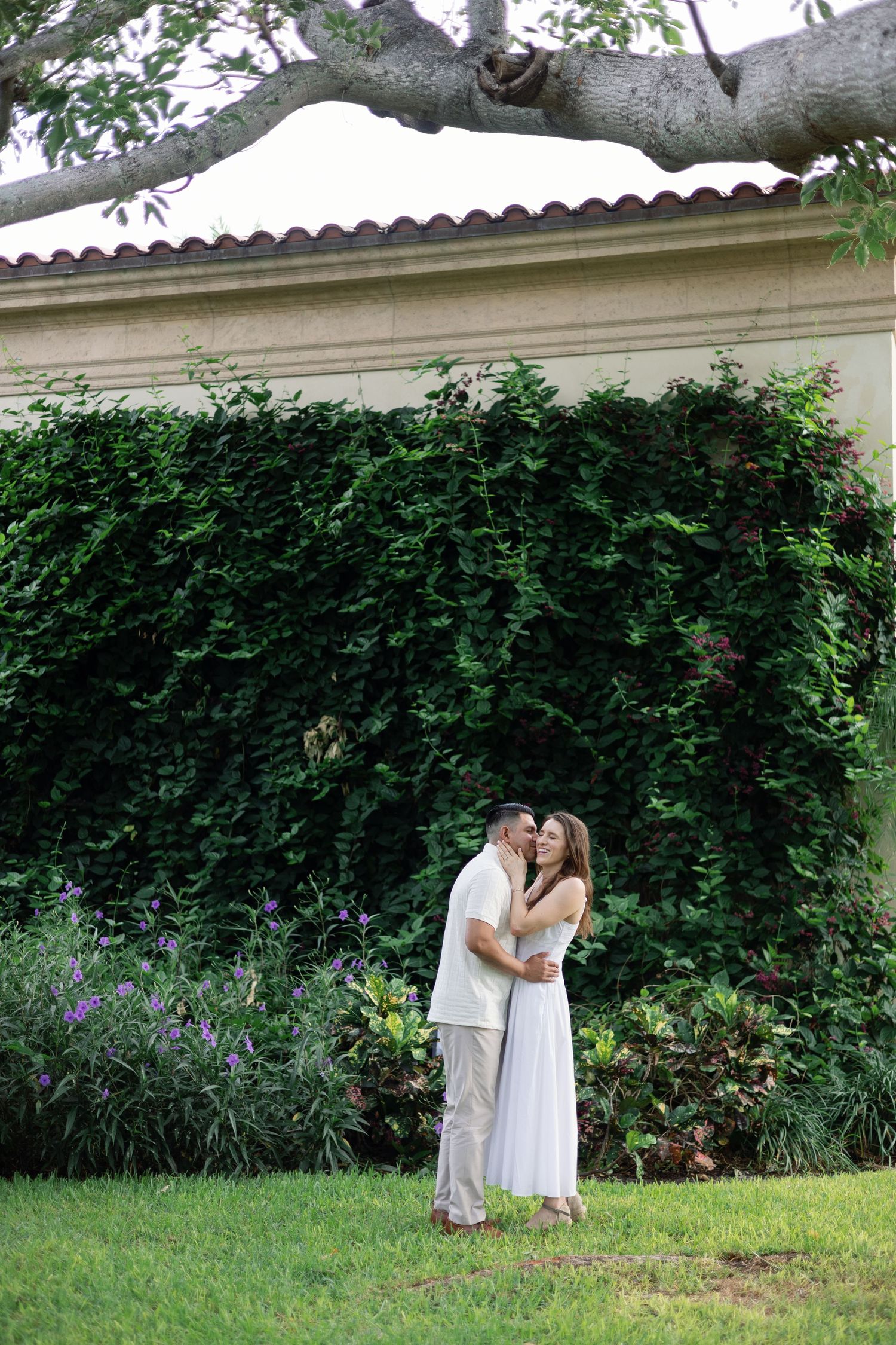 Couple embracing on green lawn before a lush ivy-covered wall and flowering garden at elegant estate.