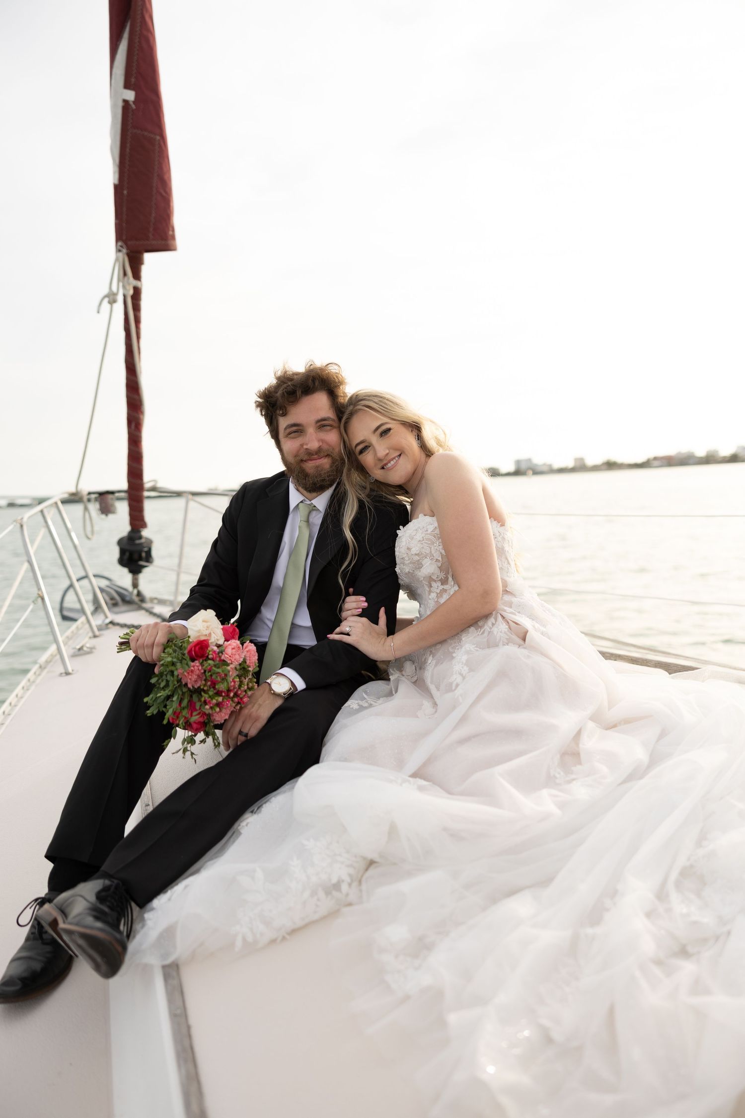 Bride and groom sitting together on the deck of a sailboat on the water holding a pink floral bouquet.
