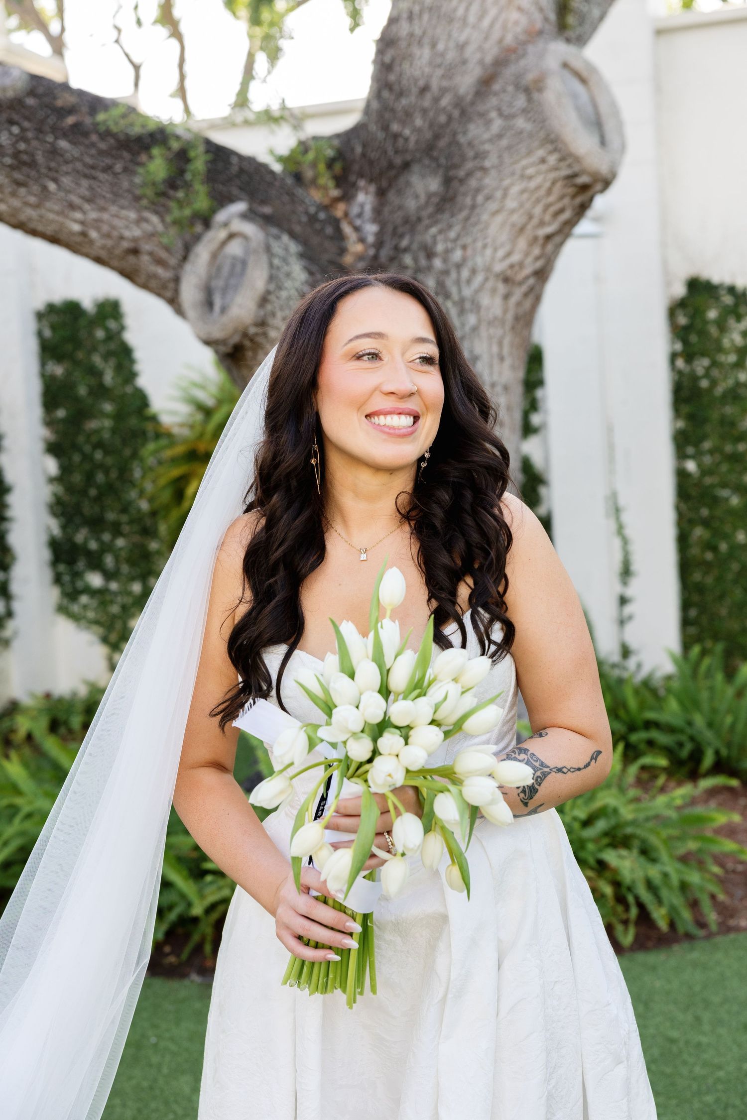 Smiling bride holding a bouquet of white tulips wearing a strapless gown and veil in a garden setting.