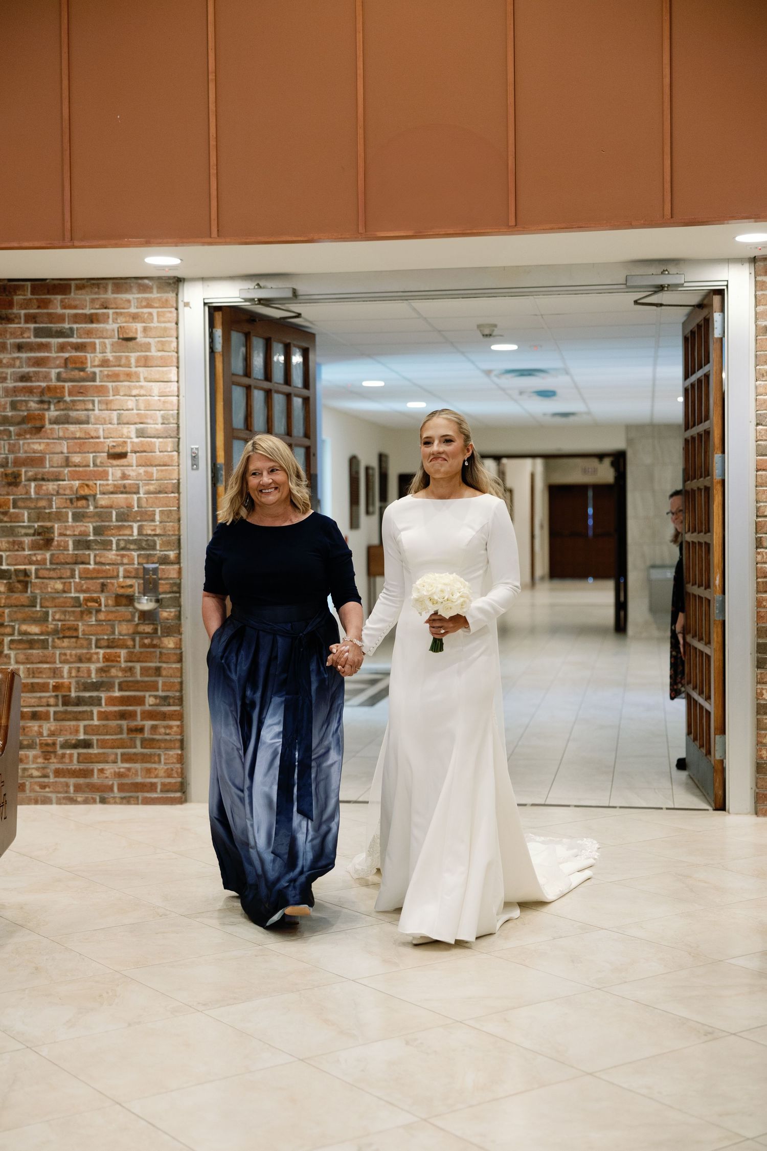 Bride in long-sleeve white gown walking with mother through church corridor holding bouquet.