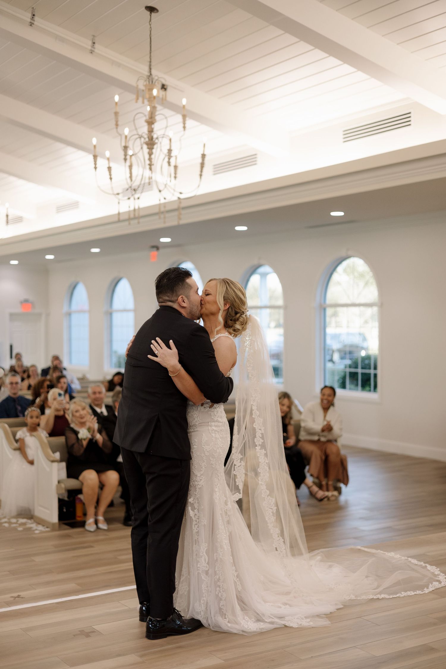 Newlyweds share first kiss at altar in bright white chapel with chandelier and arched windows.